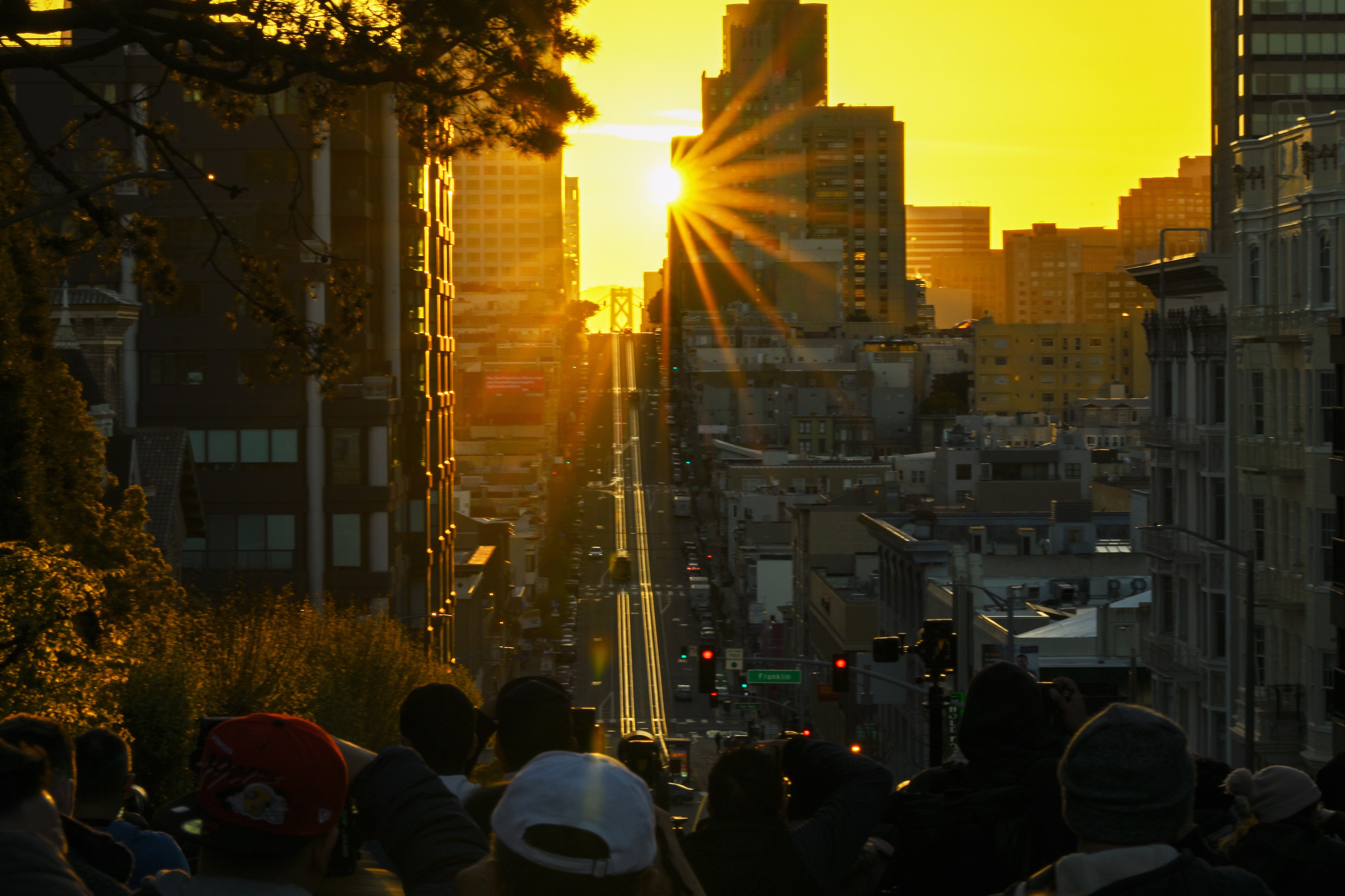 An elevated view of a city street at sunset, showing the sun's rays between buildings with silhouettes of people in the foreground.