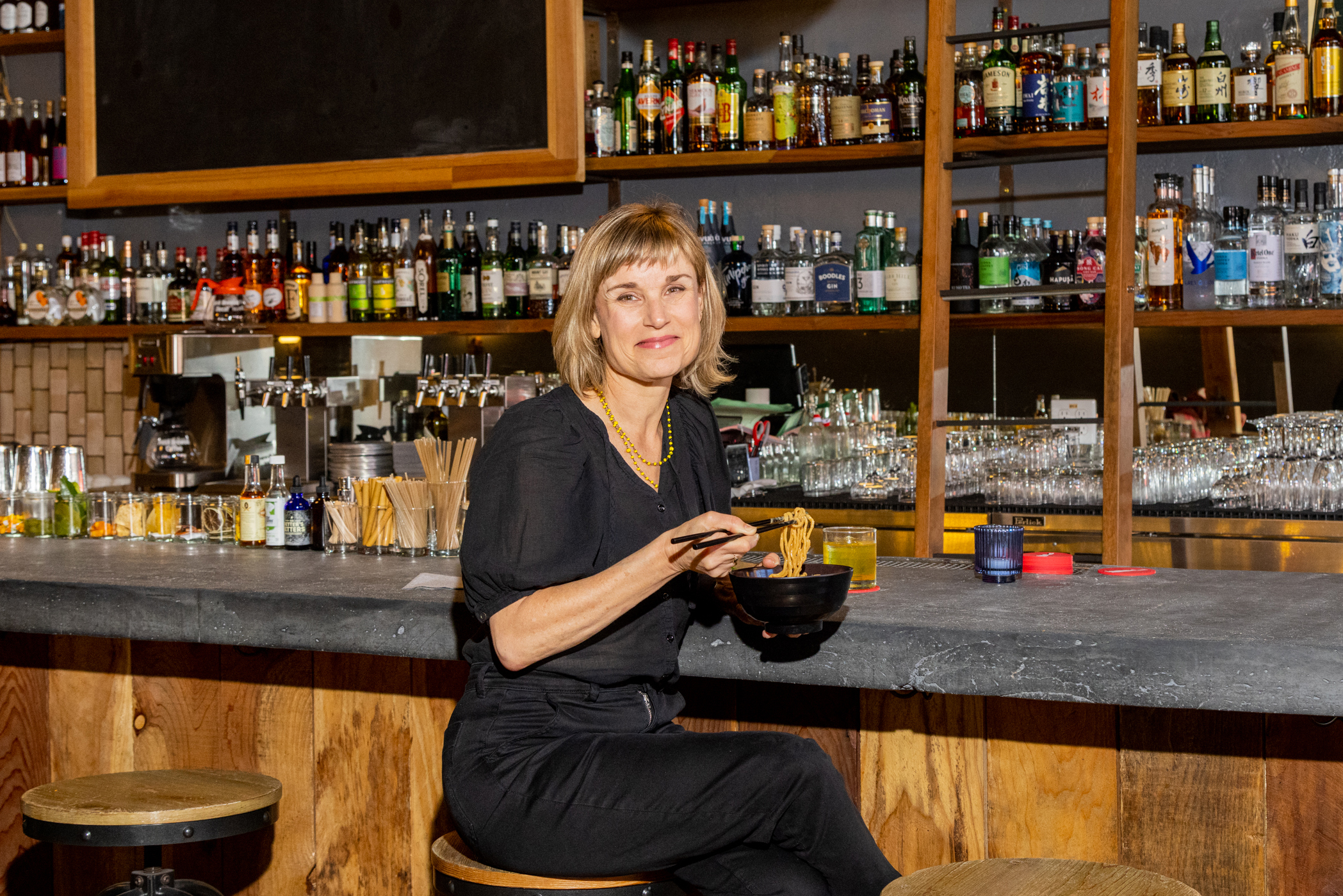A woman is smiling at a bar, holding a bowl, with shelves of liquor bottles behind her.