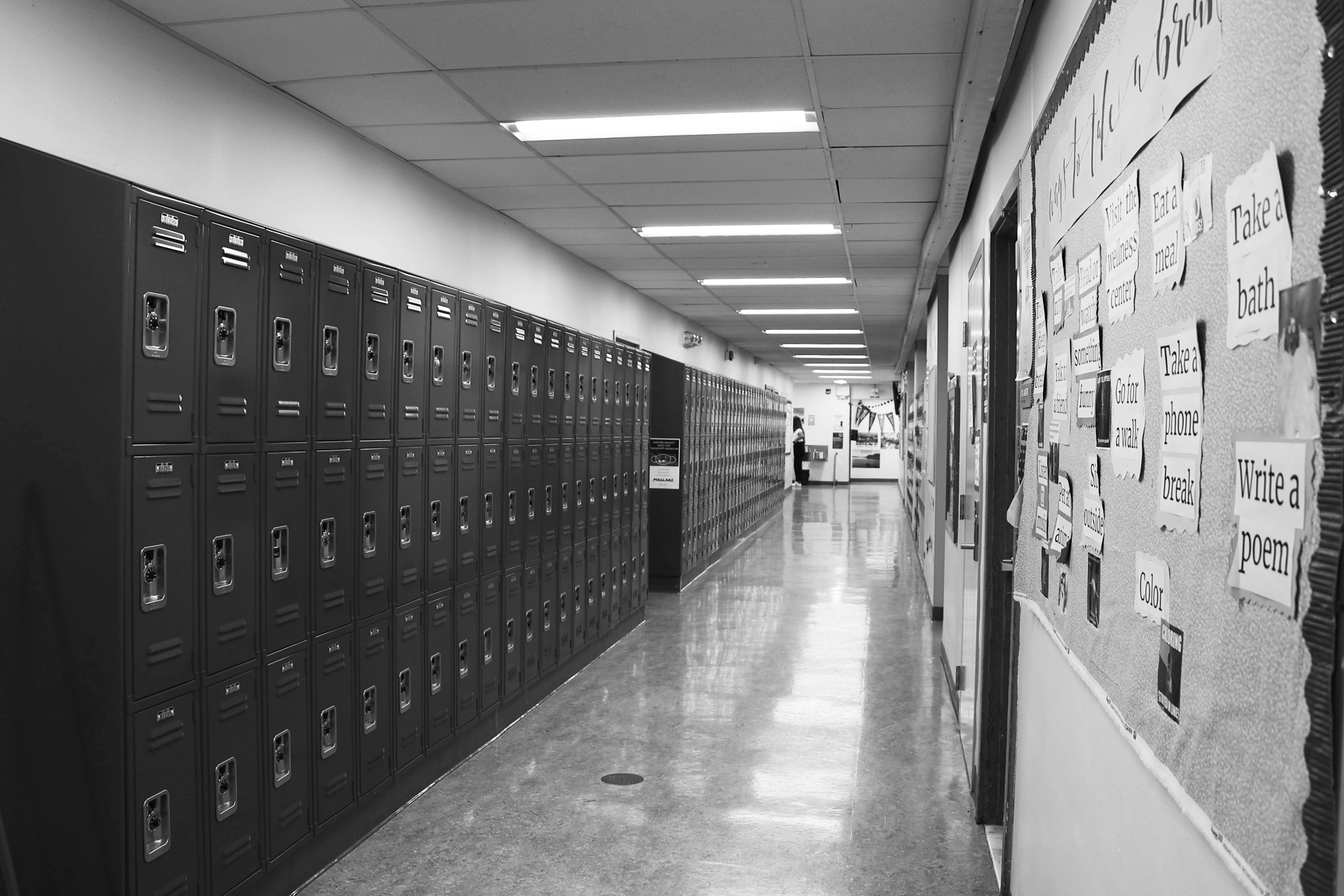 A black and white image of a long school hallway lined with rows of lockers on the left and a bulletin board on the right, under bright ceiling lights.