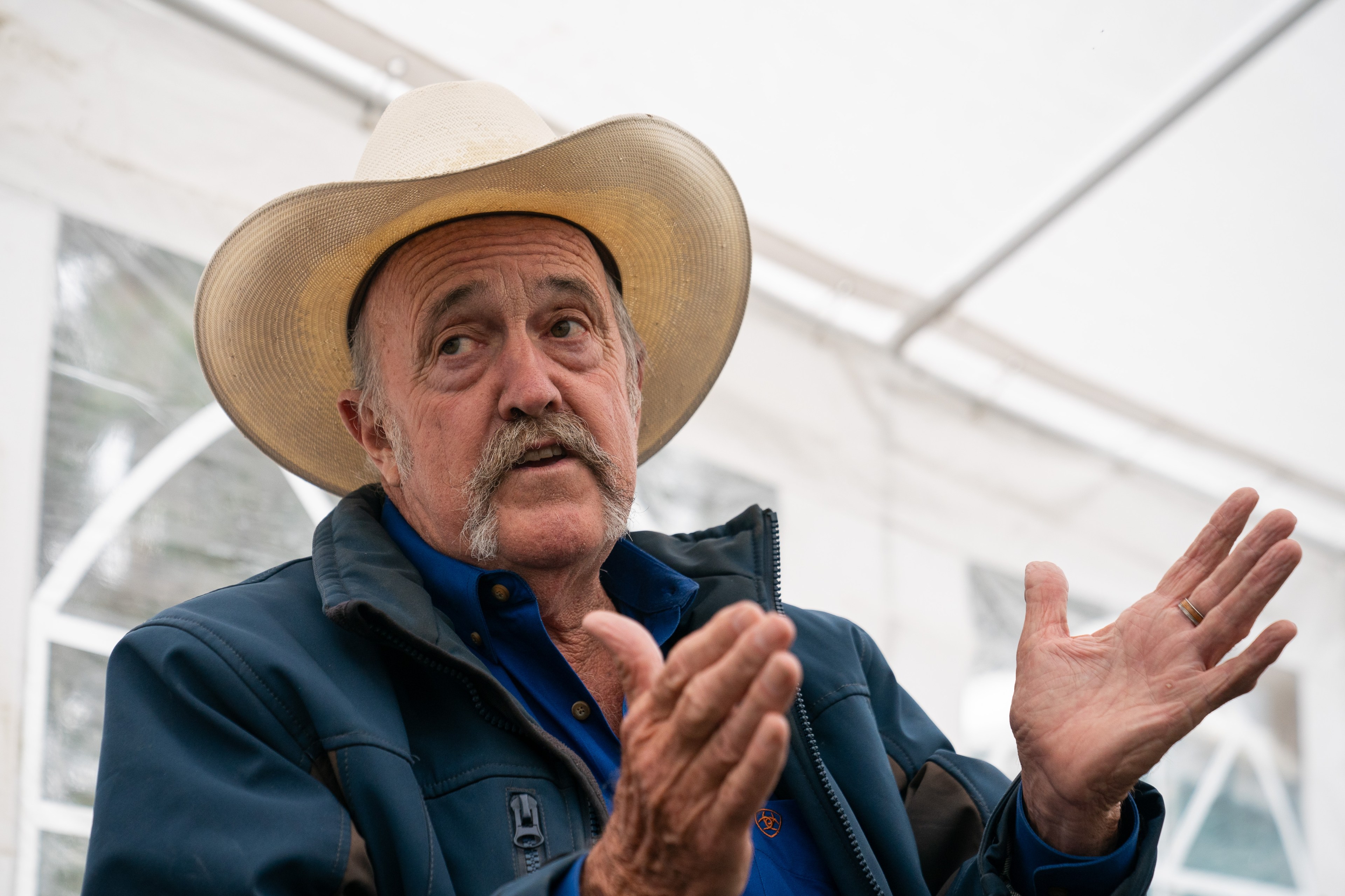 An elderly man in a cowboy hat and blue jacket is gesturing with his hands while speaking. He has a mustache and the background reveals a white tent.