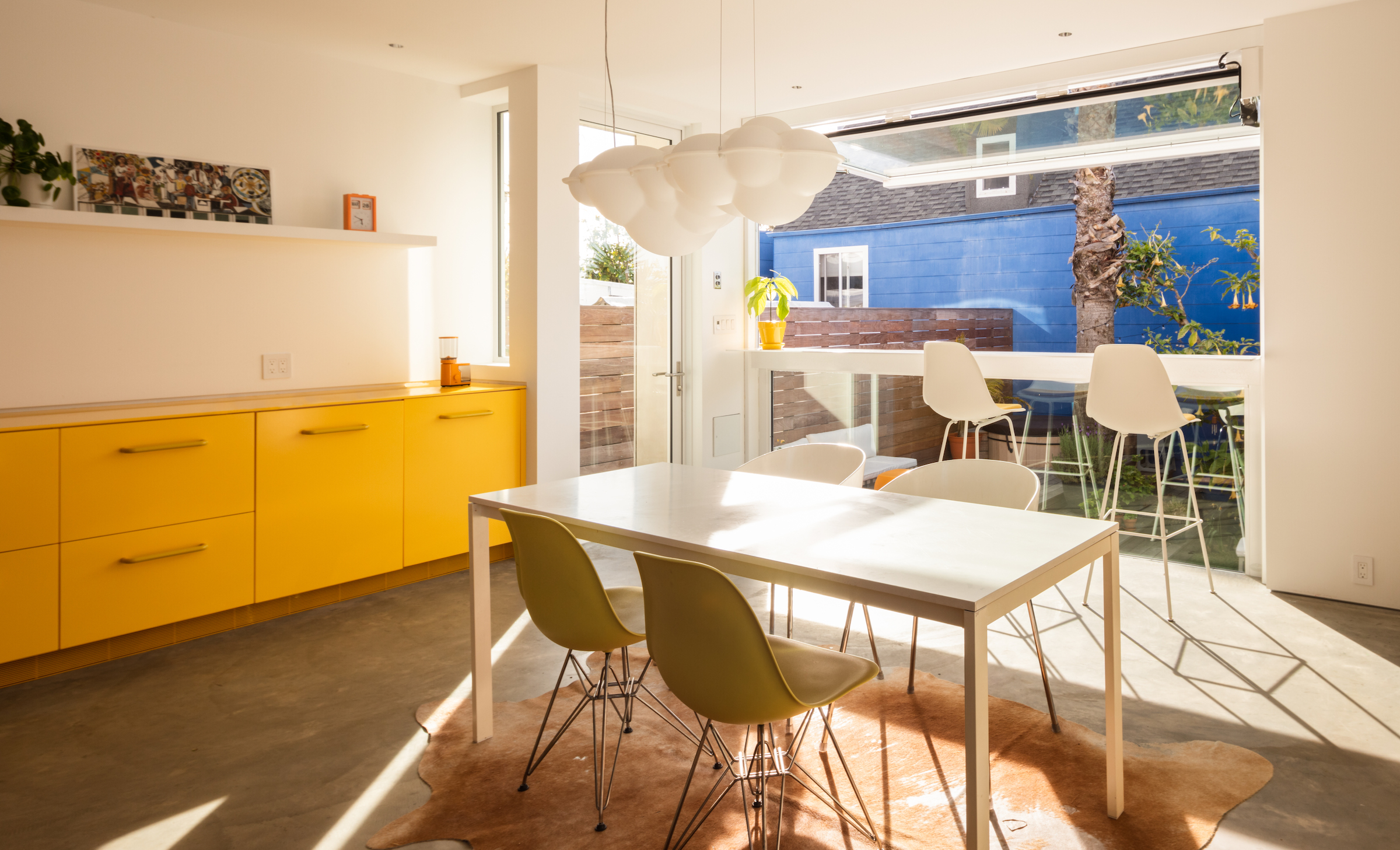 Bright dining area with a white table, olive chairs, yellow cabinets, and view to a patio with blue walls and greenery.