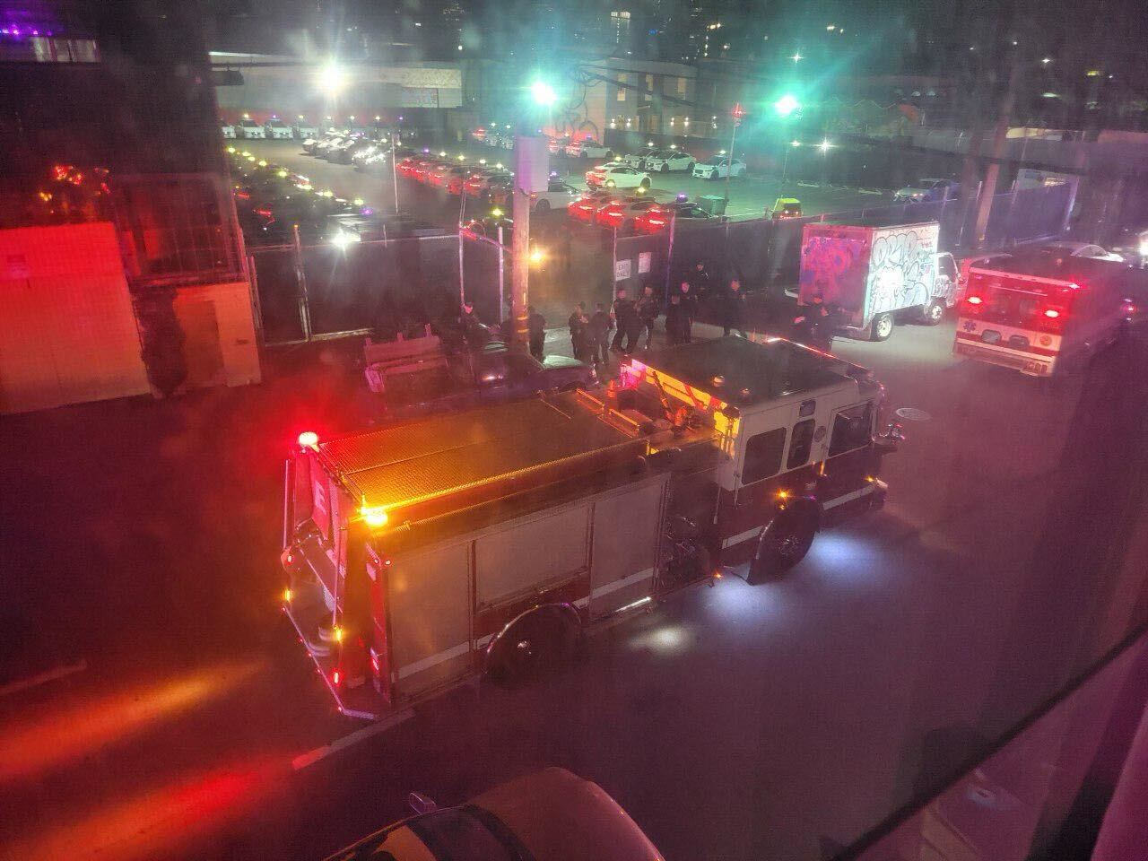 Fire trucks with flashing lights and first responders at night near a parking lot and a graffiti-covered truck.