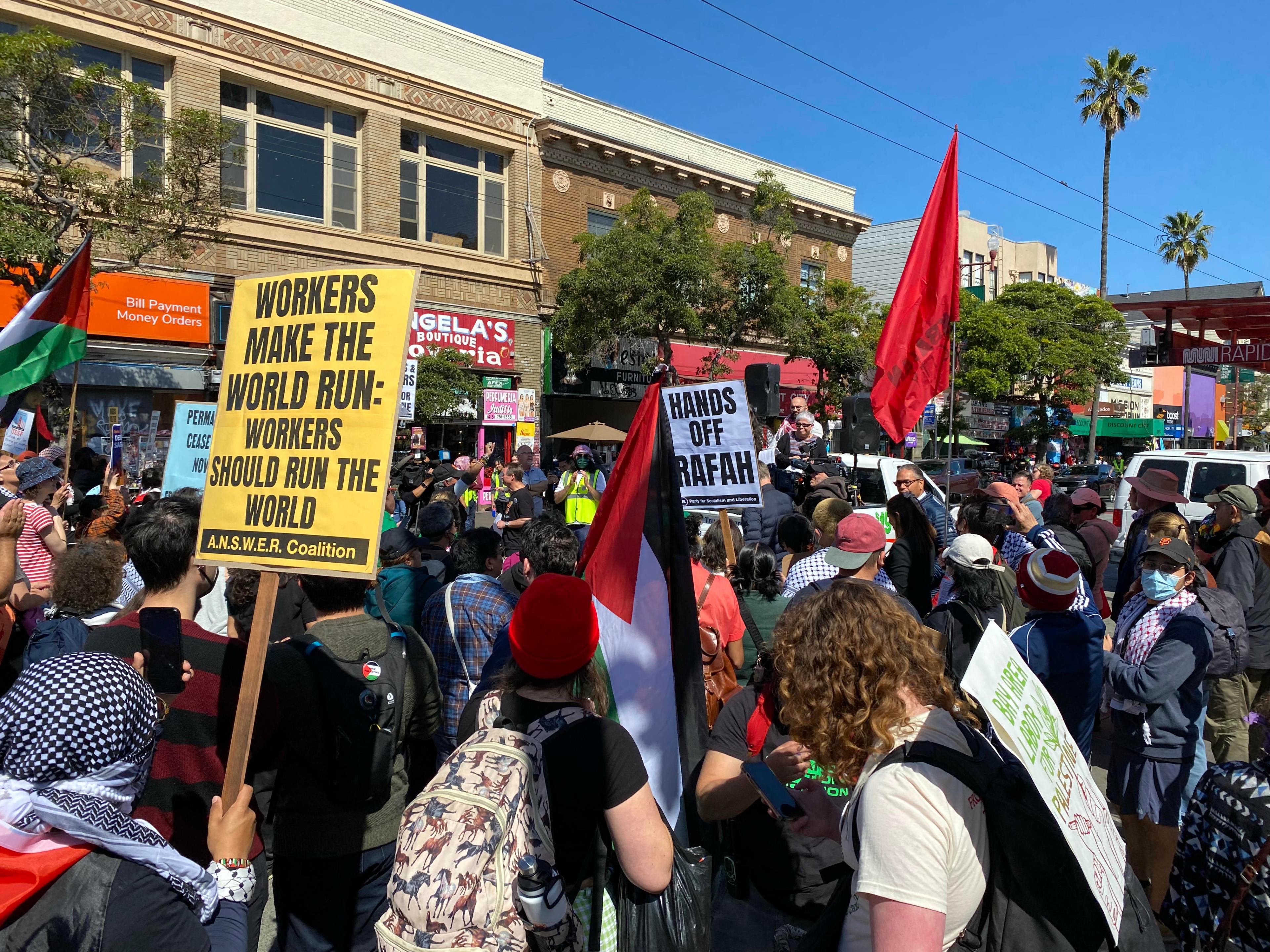 A crowd of protesters with signs, one reading &quot;WORKERS MAKE THE WORLD RUN,&quot; in an urban setting under a clear sky.