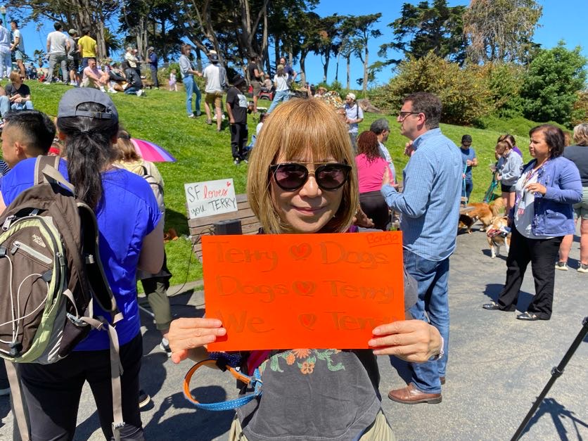 A woman with sunglasses holds a sign with hearts and &quot;Terry ❤️ Dogs&quot; in a sunny, crowded park.