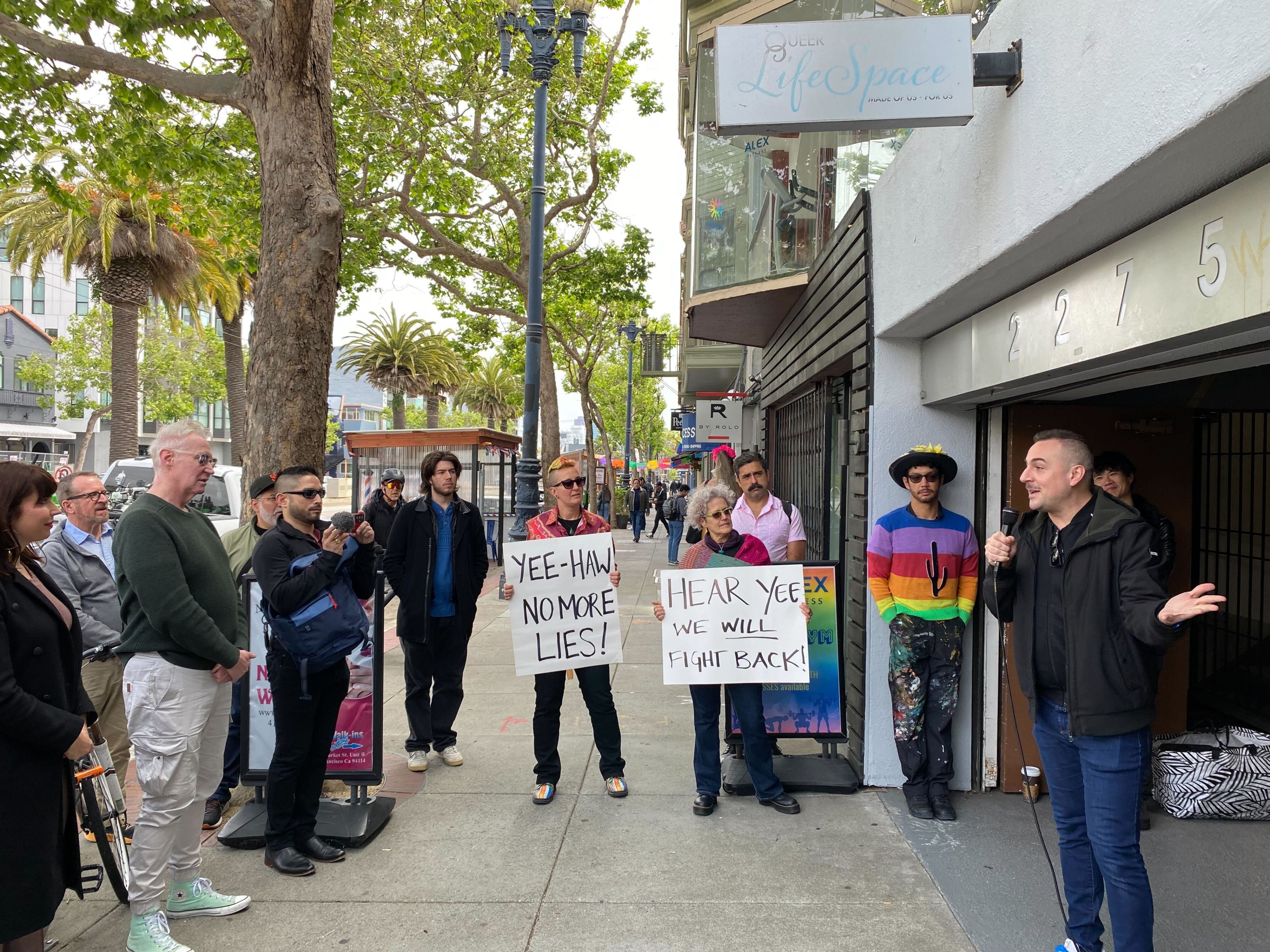 A group of people watches a man speaking into a microphone on a city sidewalk, flanked by two holding protest signs with slogans about truth.