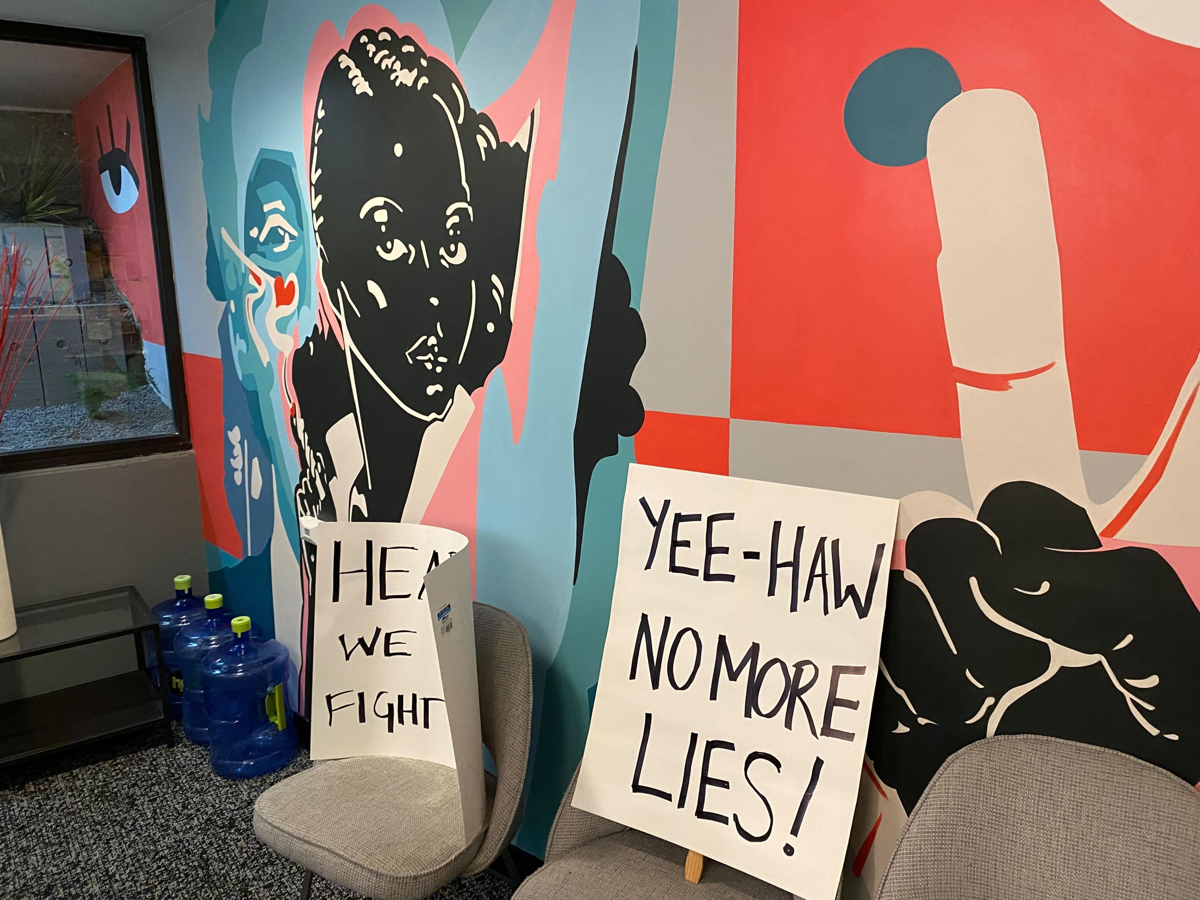 A colorful mural with abstract figures and a dog's face, has protest signs reading &quot;HEAR WE FIGHT&quot; and &quot;YEE-HAW NO MORE LIES!&quot; placed near comfy chairs.