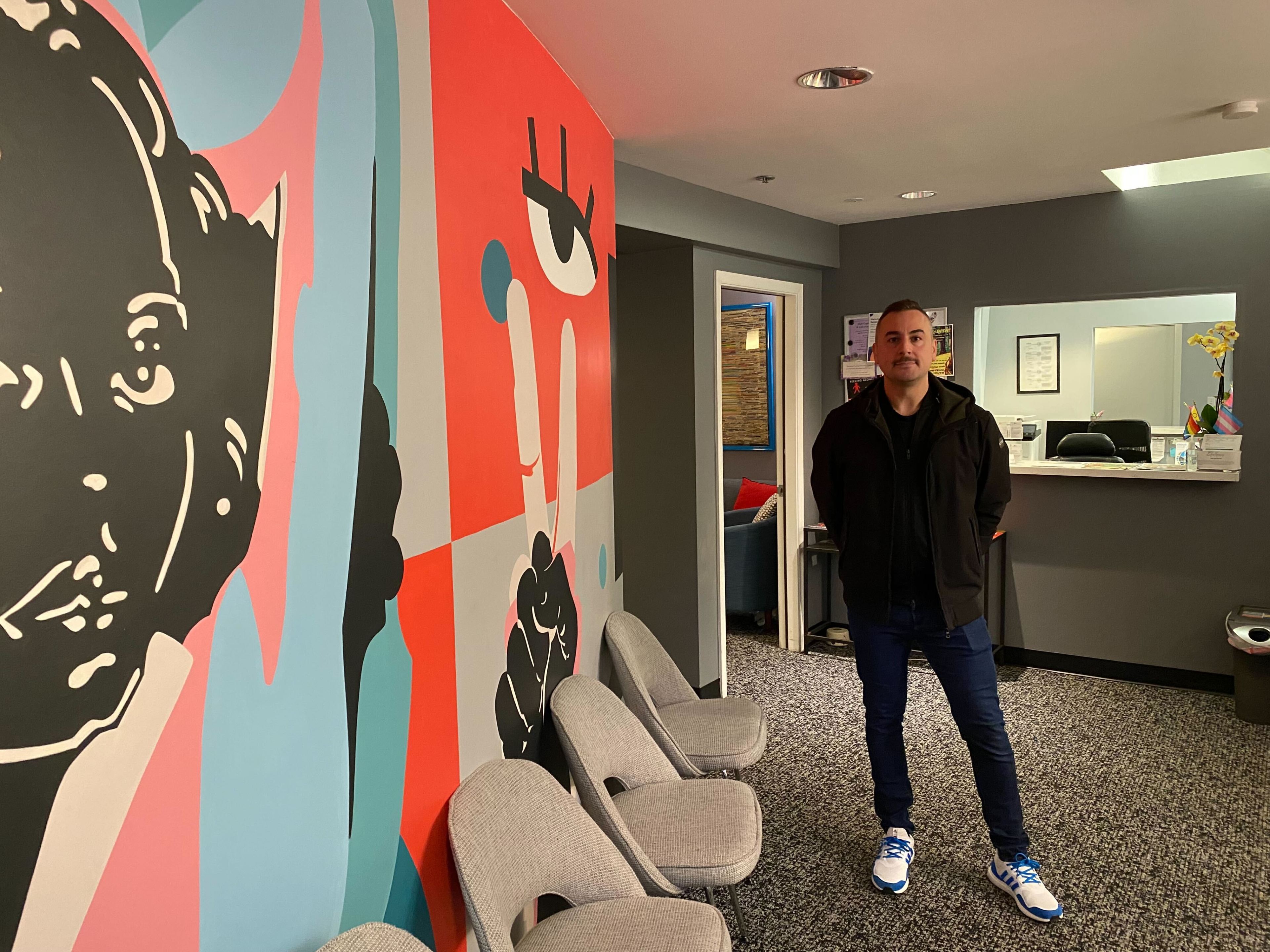 A man stands in an office with large colorful abstract wall art, wearing a black jacket, blue jeans, and white shoes.