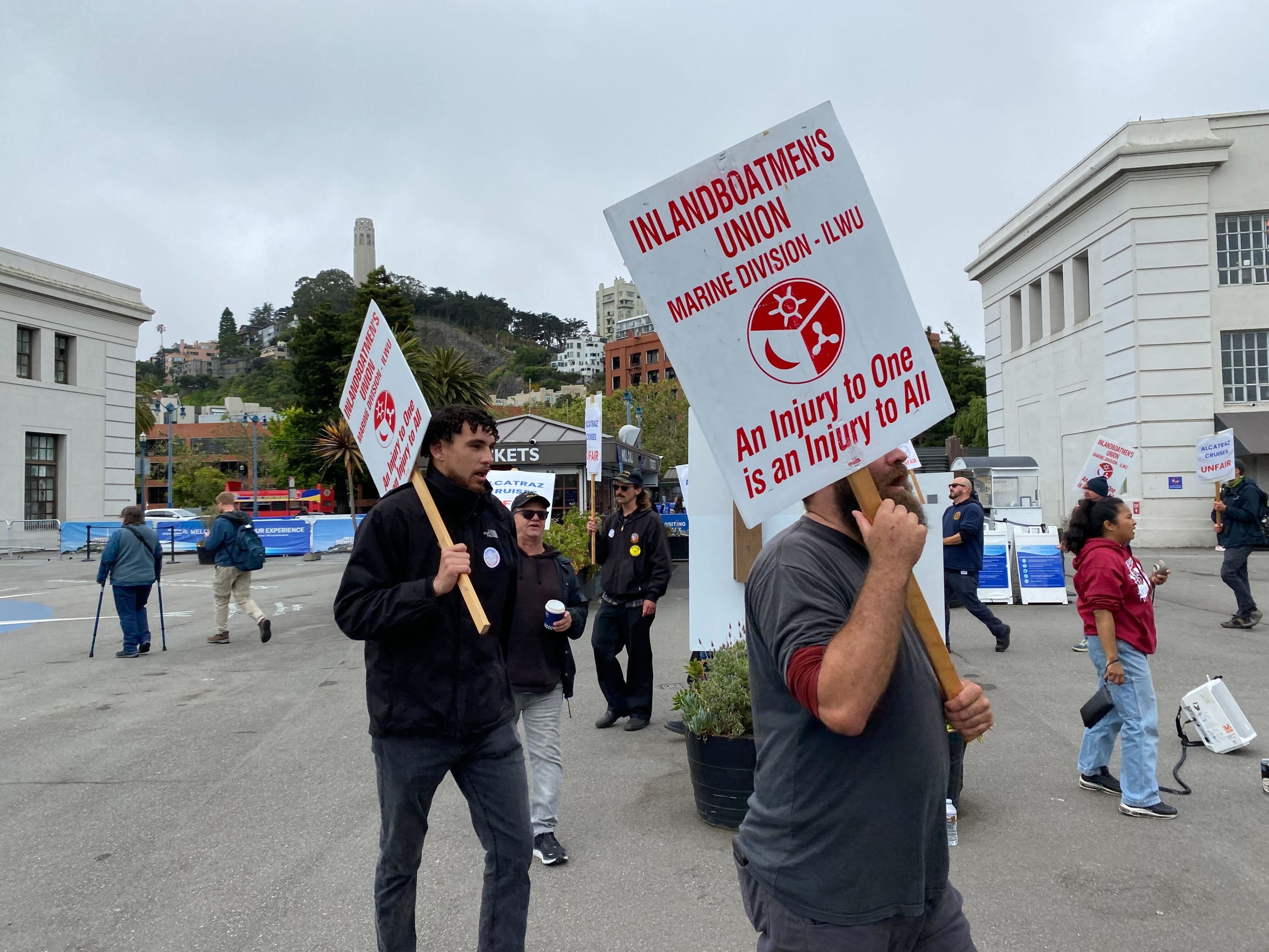 People are protesting outdoors, holding signs that read, "Inlandboatmen's Union, Marine Division - ILWU. An Injury to One is an Injury to All." A tall structure and buildings are in the background.