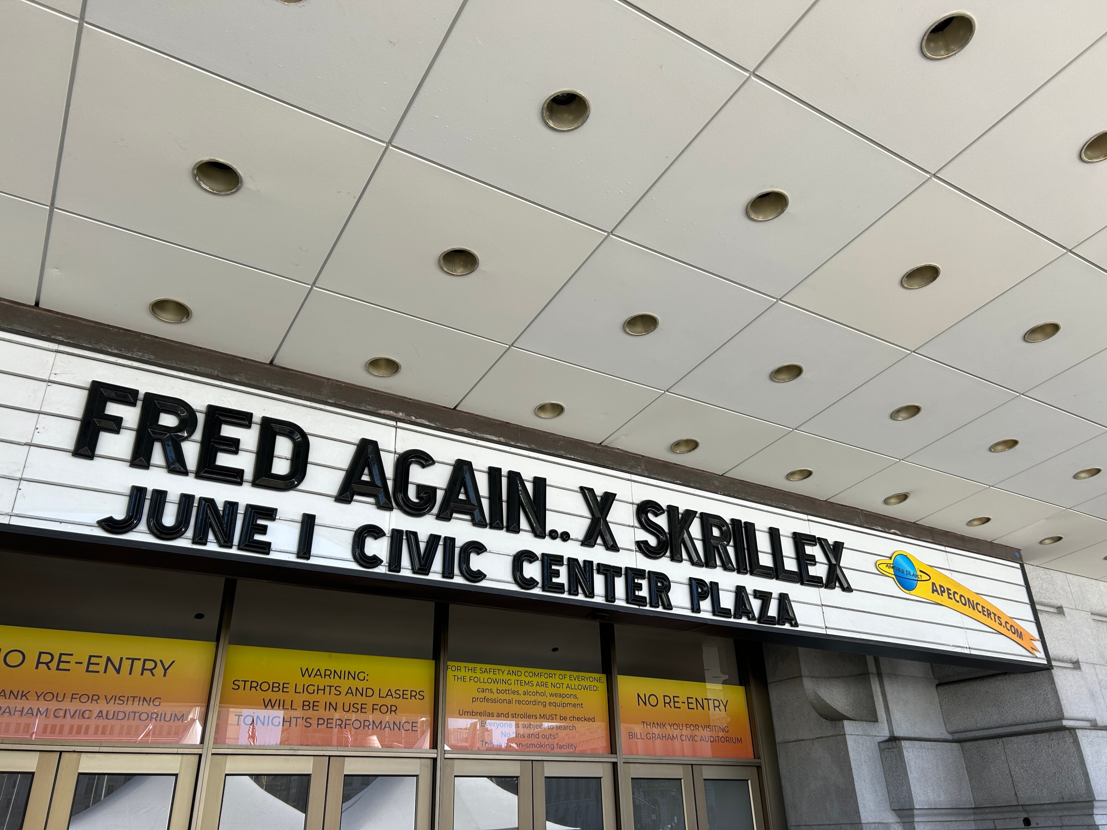 A marquee reads &quot;FRED AGAIN.. X SKRILLEX JUNE 1 CIVIC CENTER PLAZA.&quot; Below are doors and signs about re-entry, strobe lights, and safety information.