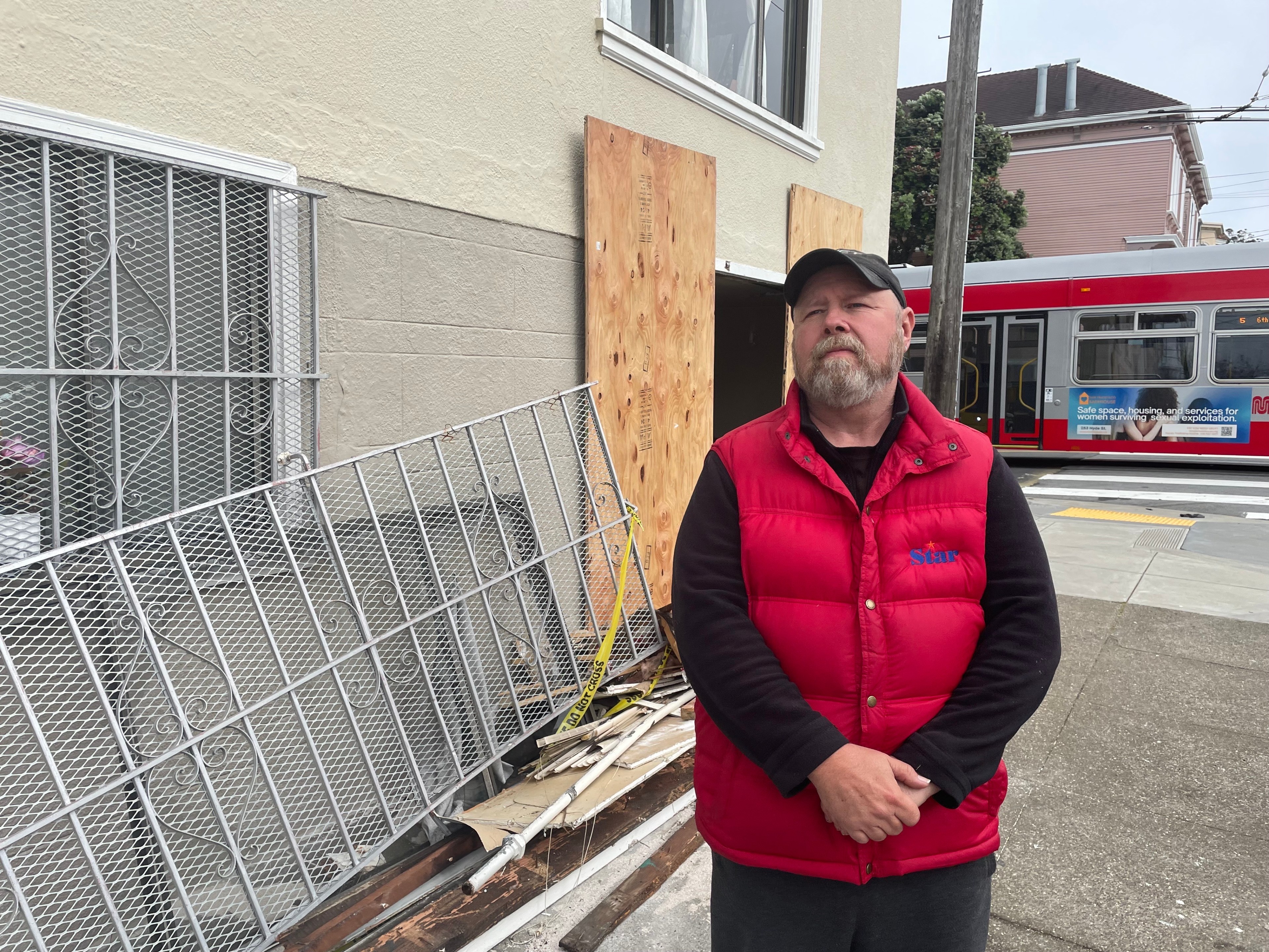 A man in a red vest stands in front of a building with boarded-up windows, near a metal fence and red trolley bus in the background.