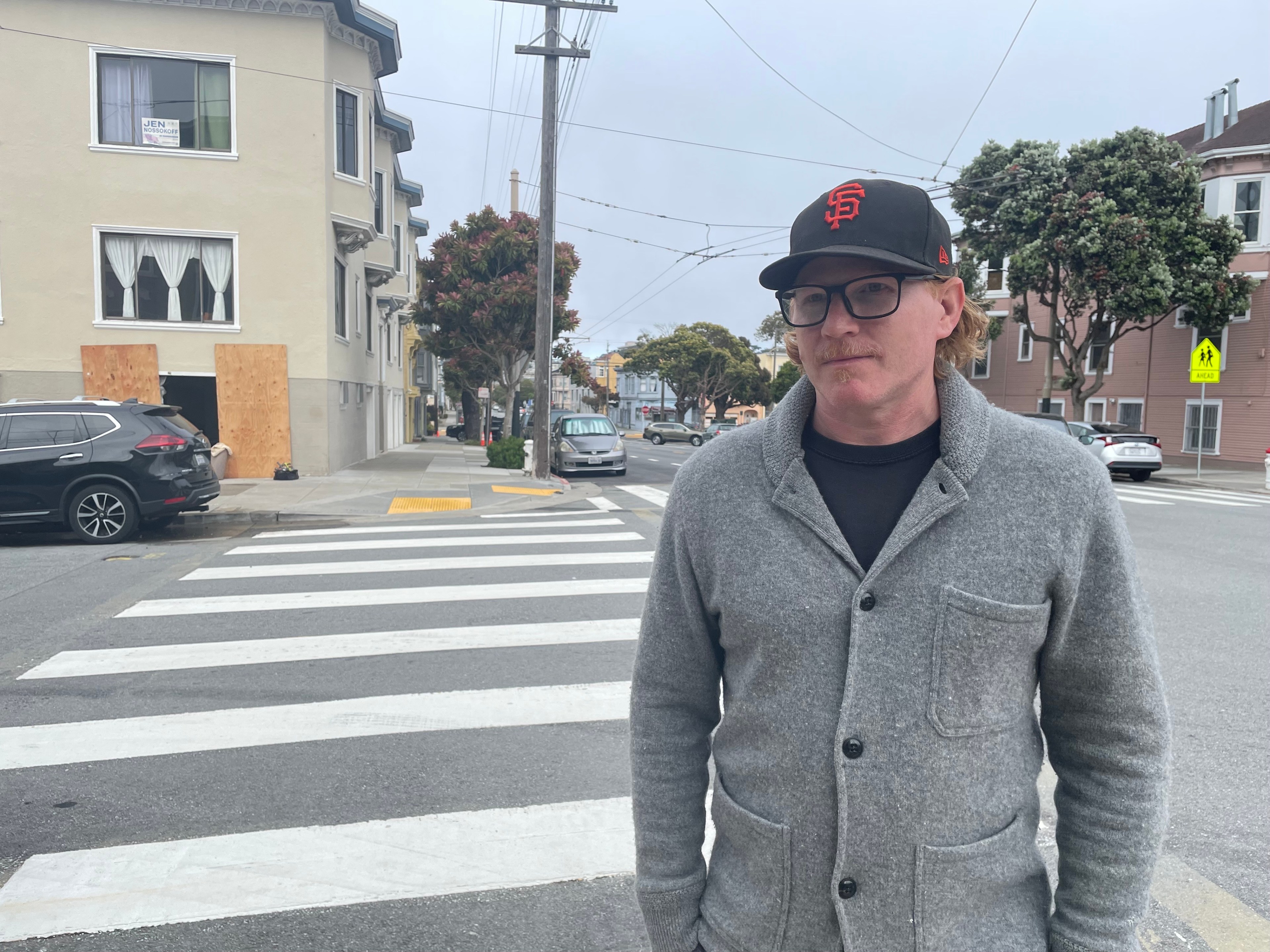 A man stands before a crosswalk on a city street, wearing a grey coat and a baseball cap with the SF Giants logo. The neighborhood has residential buildings and parked cars.