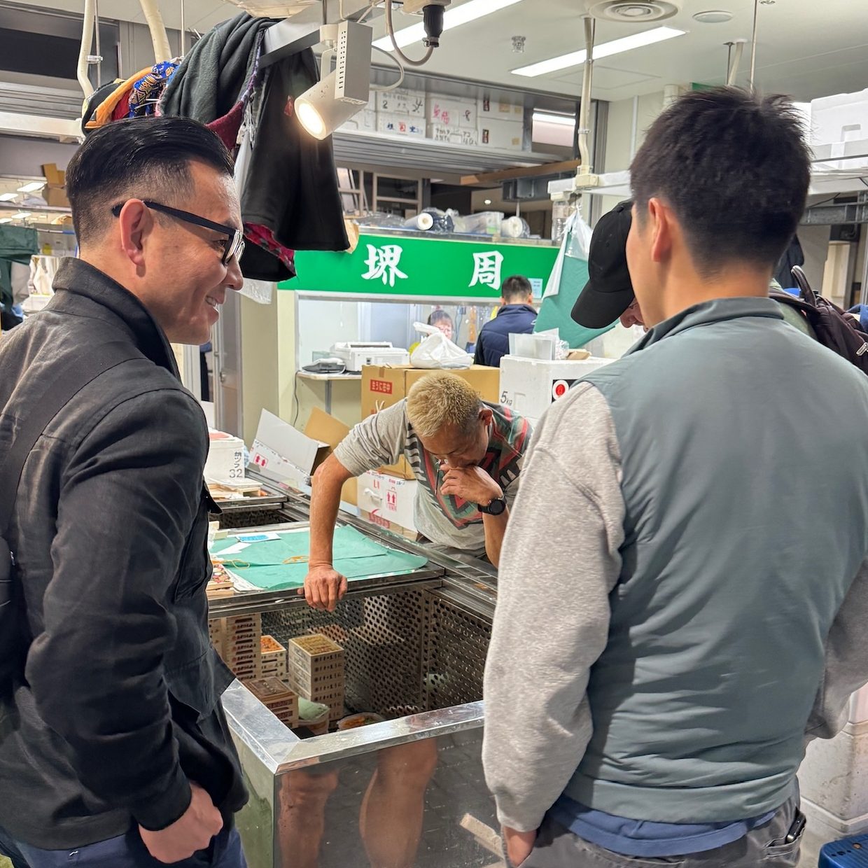 Two men are talking to a fish vendor in an indoor market. The vendor is leaning over a tank, while the men observe. The area is well-lit and busy.