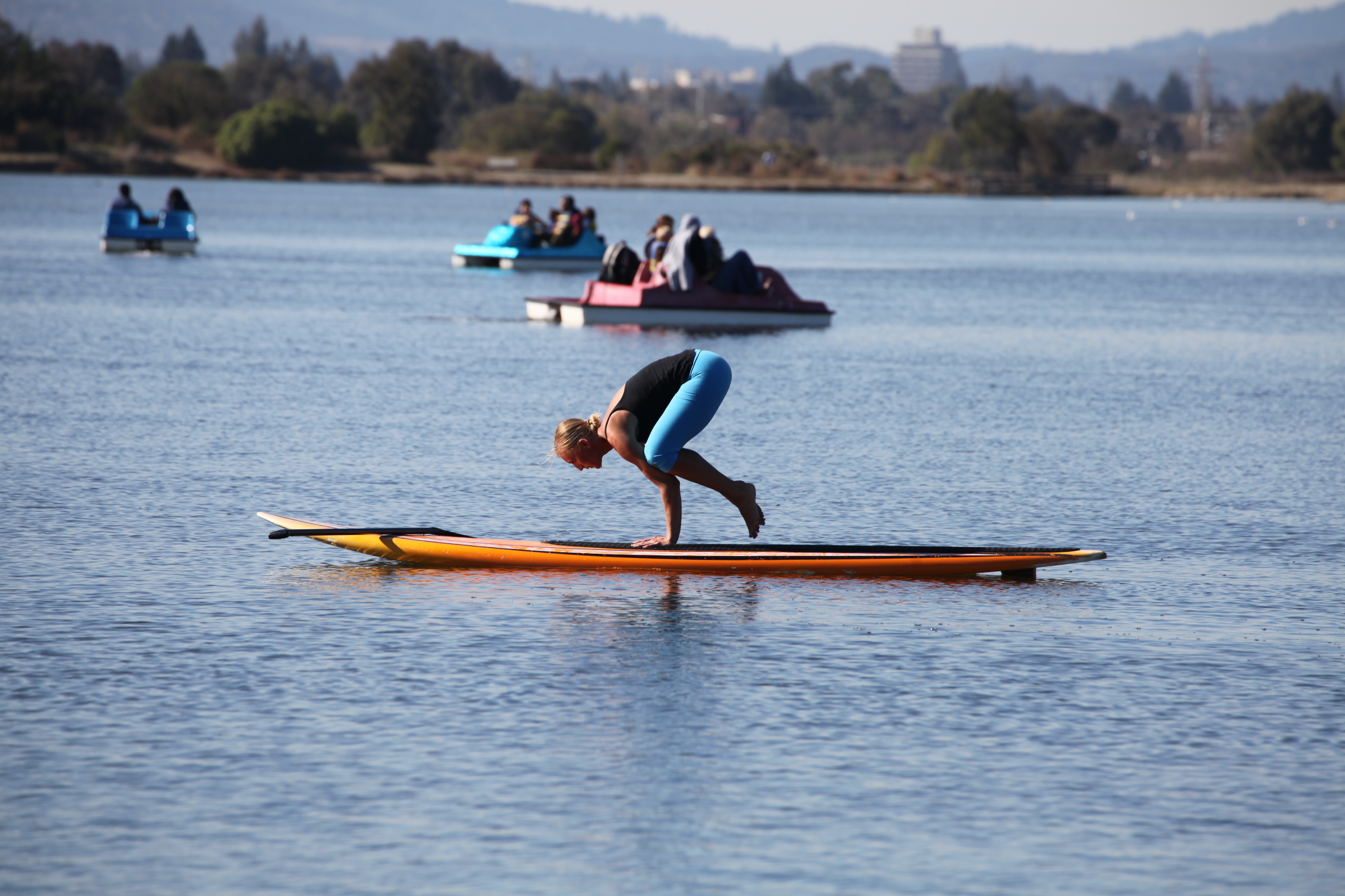 A person in a wetsuit balances on a paddleboard, with pedal boats in the background on calm water.