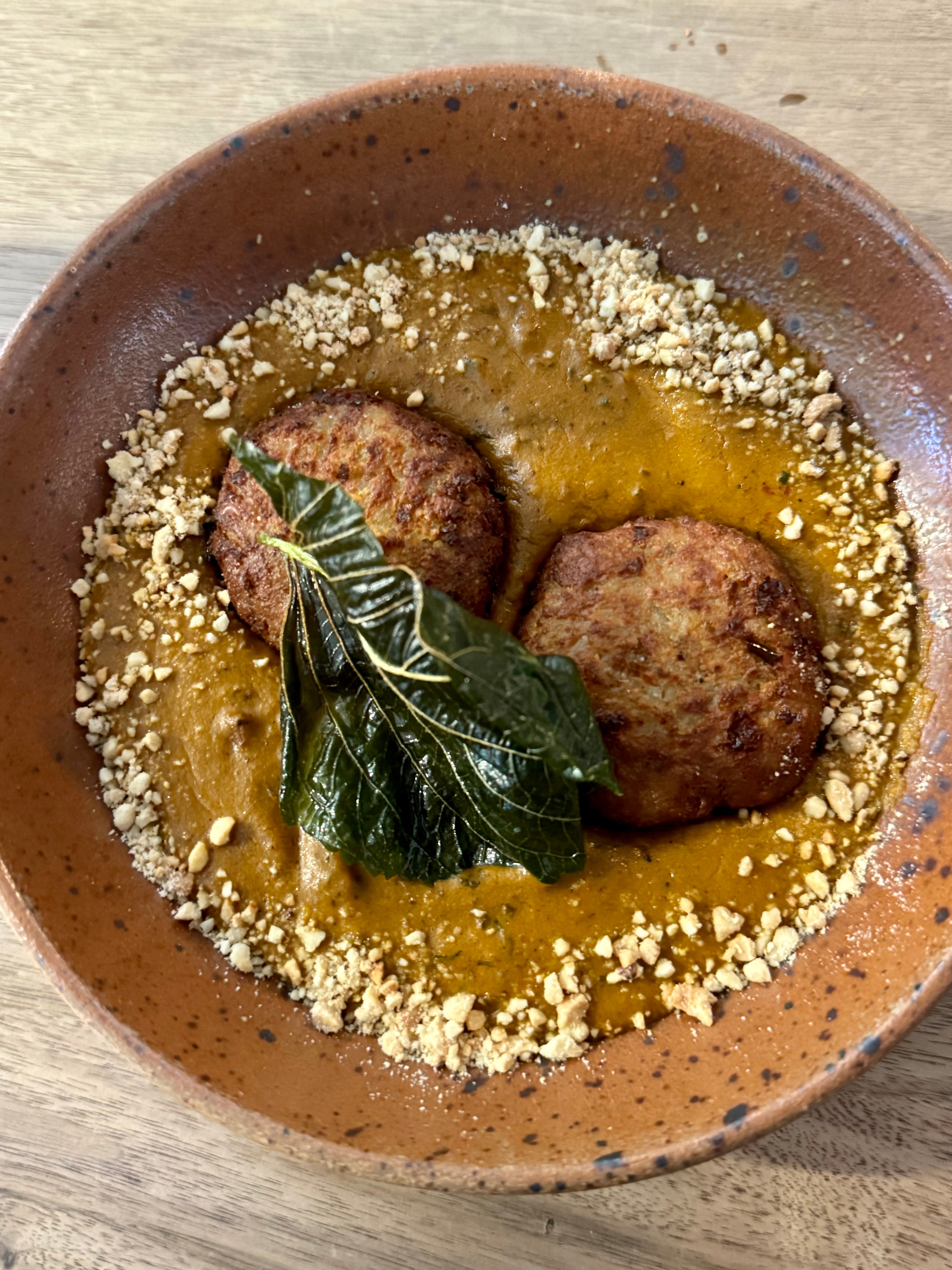 A bowl of food with two croquettes, a brown-orange cashew sauce, crushed nuts, and a leaf garnish.