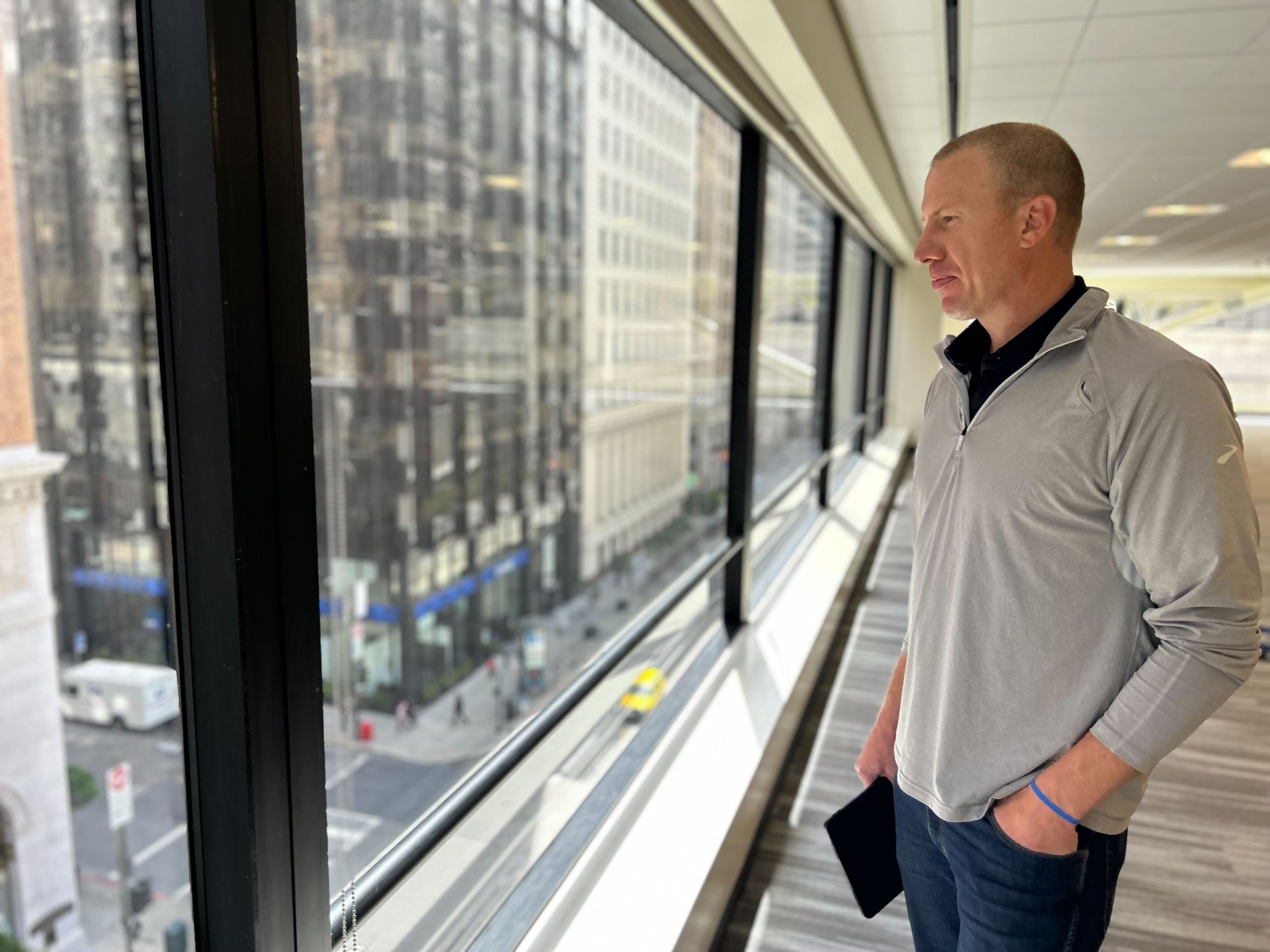 A man in a gray shirt stands by large windows overlooking a busy city street, holding a tablet and gazing outward thoughtfully.