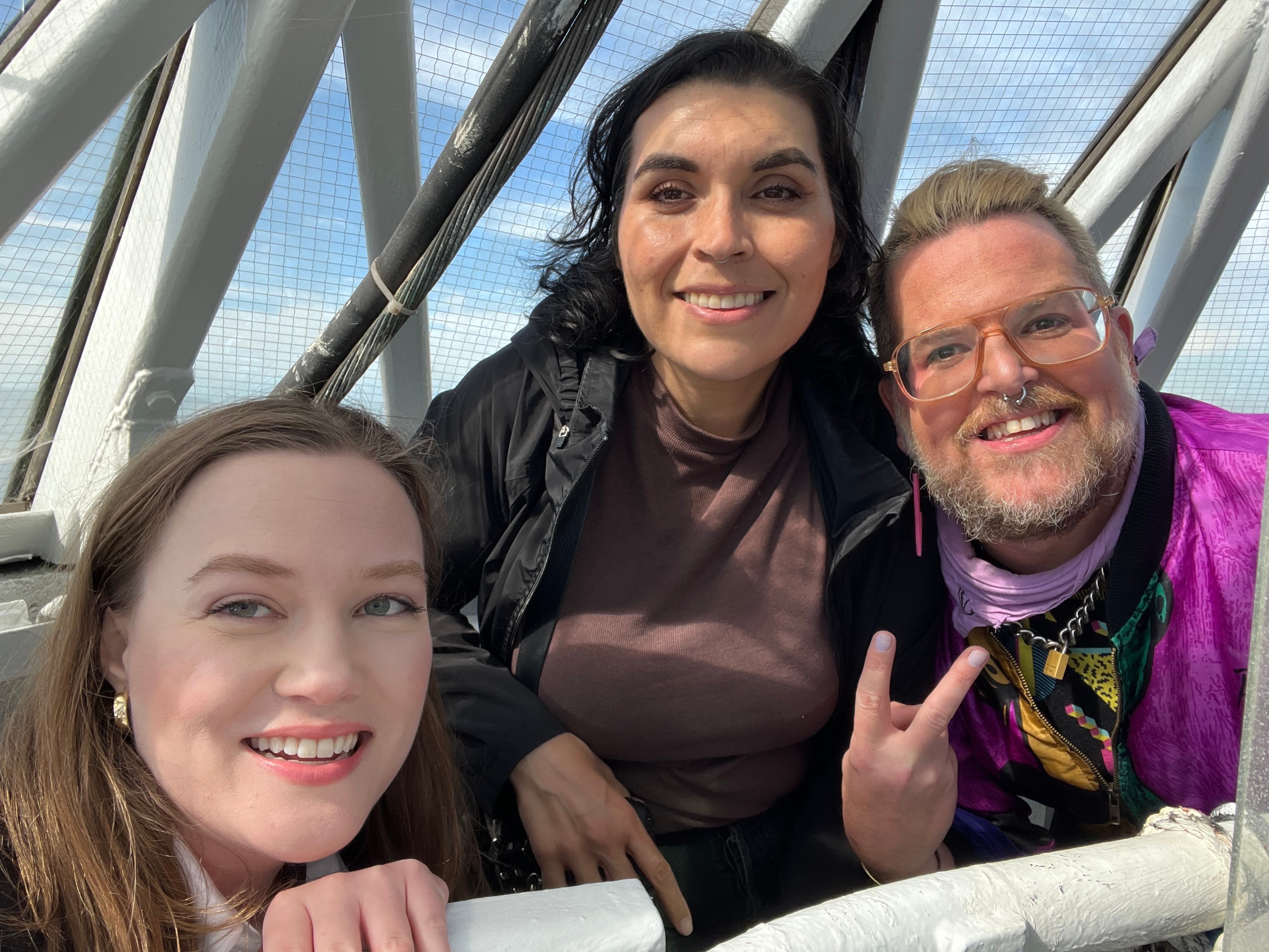 Three smiling people are taking a selfie, with metal beams and netting in the background, suggesting an outdoor setting.