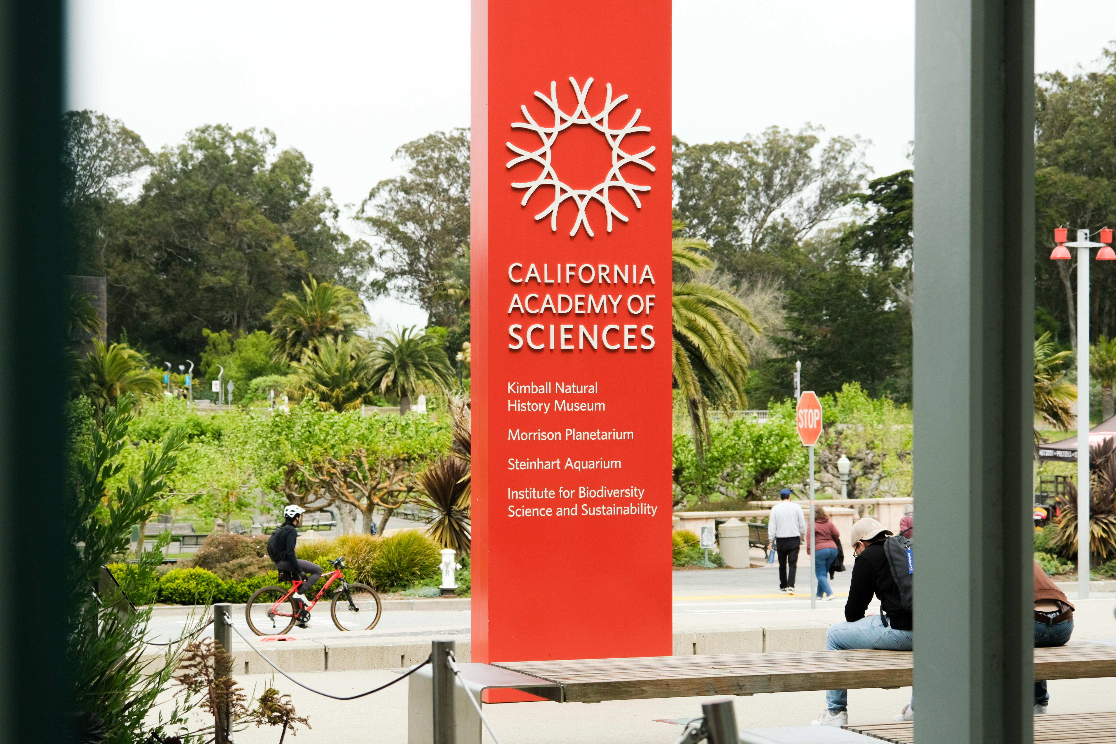 A bright red sign with &quot;California Academy of Sciences&quot; stands before lush greenery, listing museum attractions. Cyclists and pedestrians are nearby.