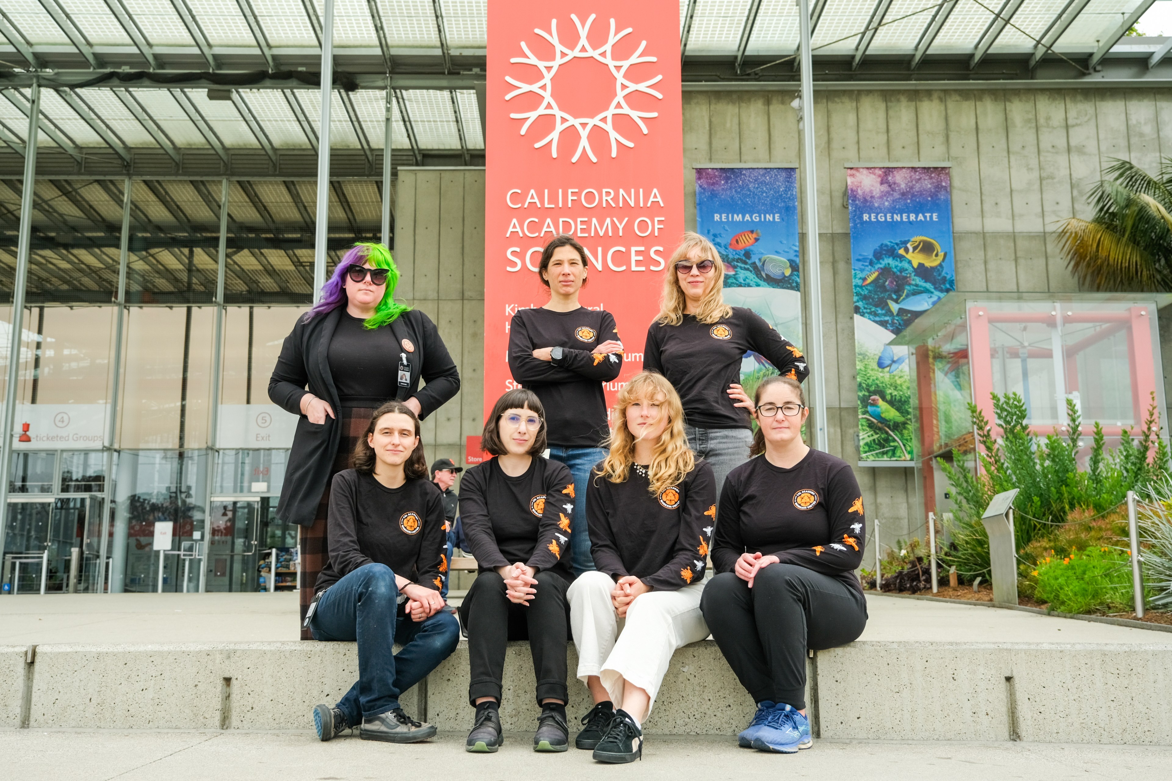 Seven people in matching black tops with orange logos pose in front of a California Academy of Sciences sign, exuding a team spirit.