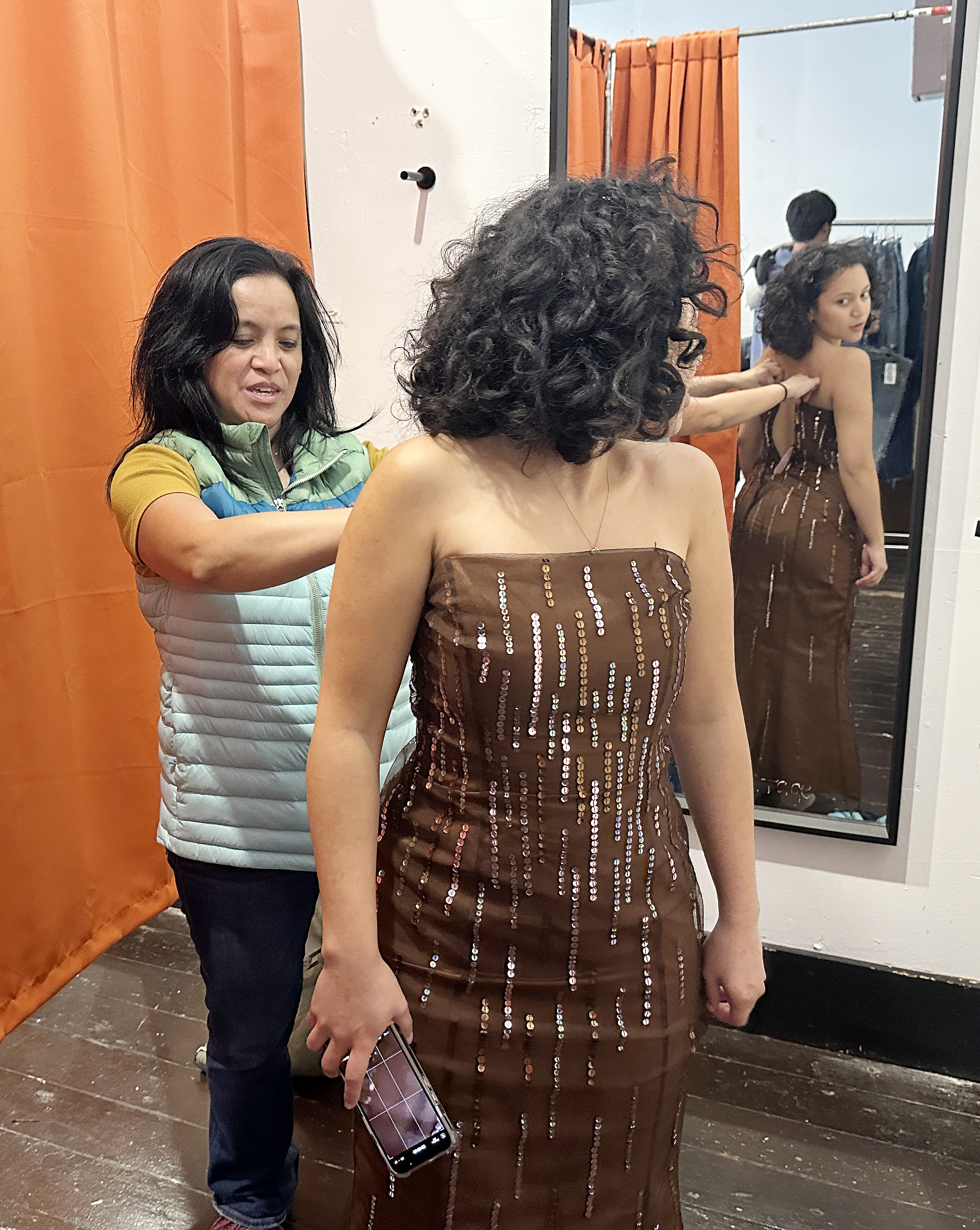 A woman is assisting another in fitting a sequined dress, viewed in a mirror within a dressing room.