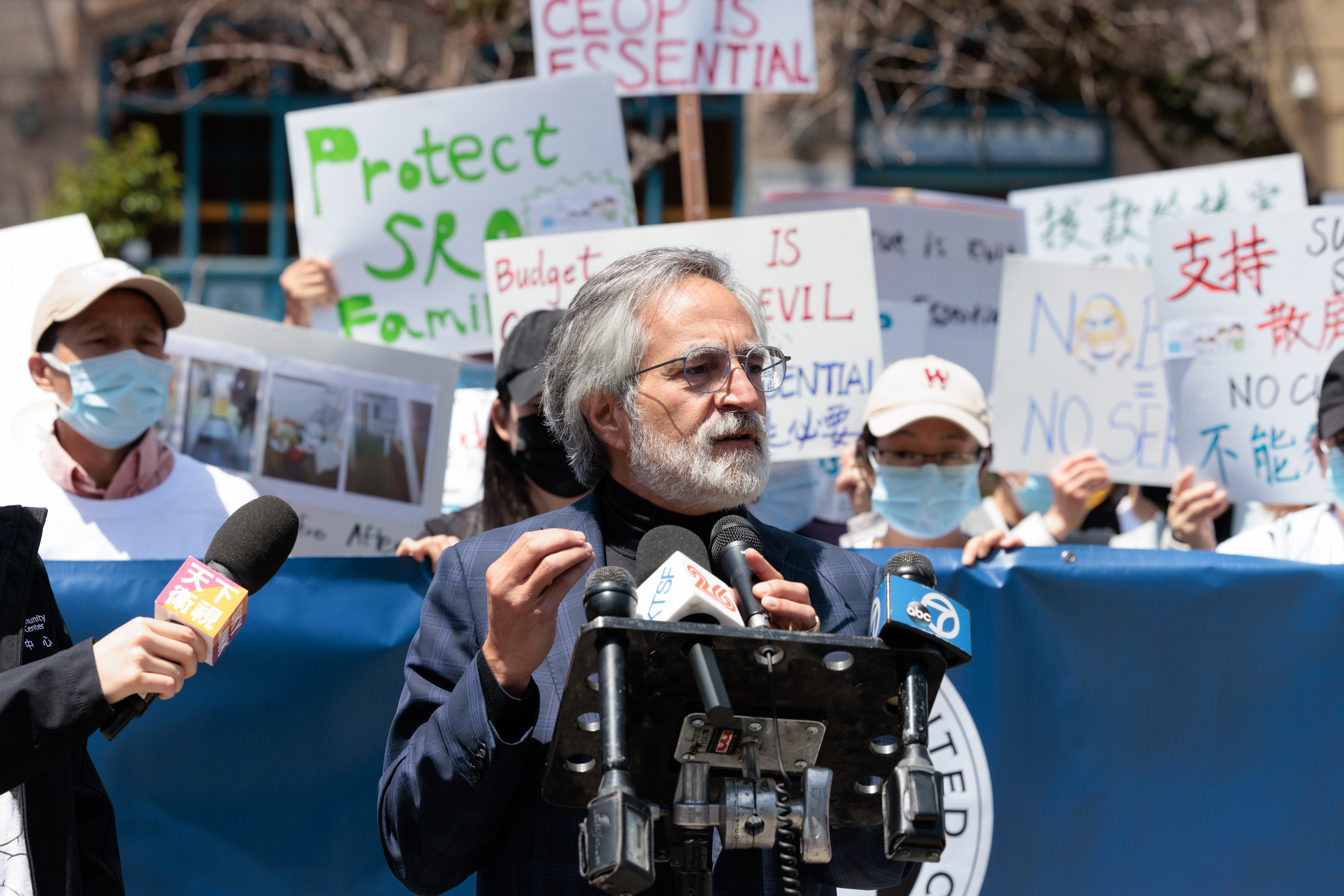 A man speaks at a podium with multiple microphones, surrounded by people holding protest signs in various languages. Some wear masks and hats.