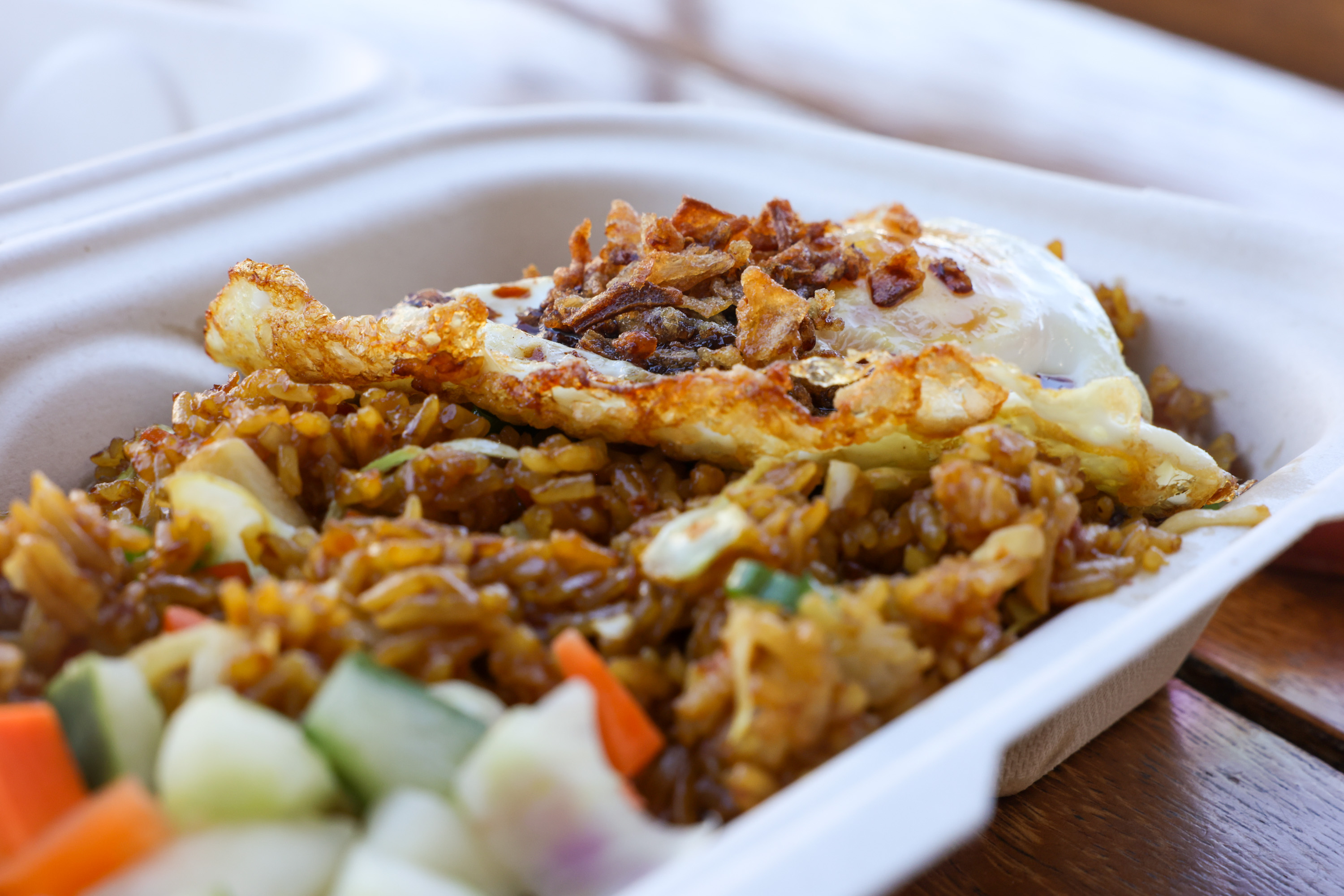Fried rice topped with a sunny side up egg and crispy fried onions, served with a side of diced cucumber and carrots in a white takeout container.