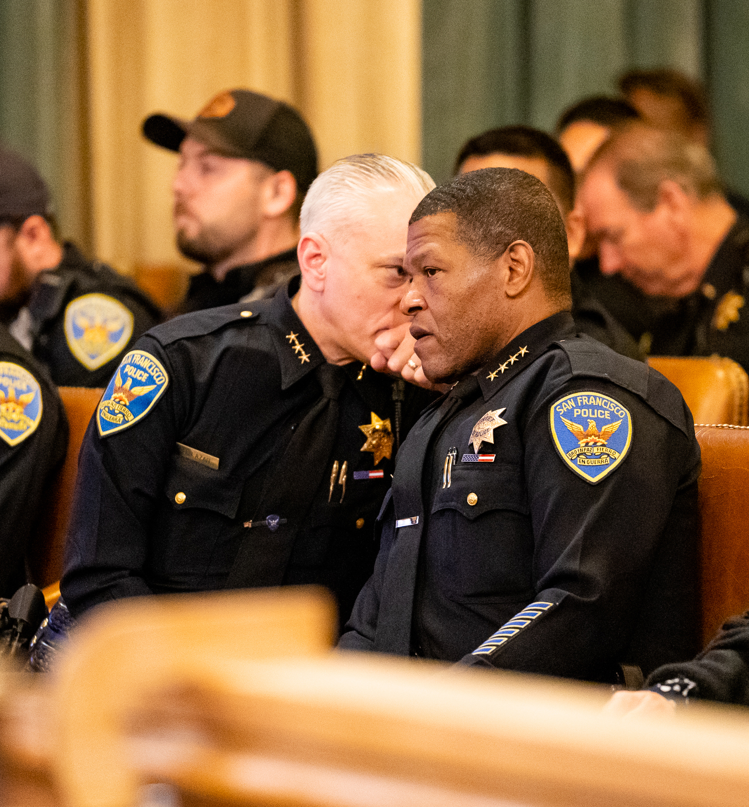 Two police officers in uniform sitting closely and conversing in the Board of Supervisors chambers, with several other uniformed officers seated in the background.