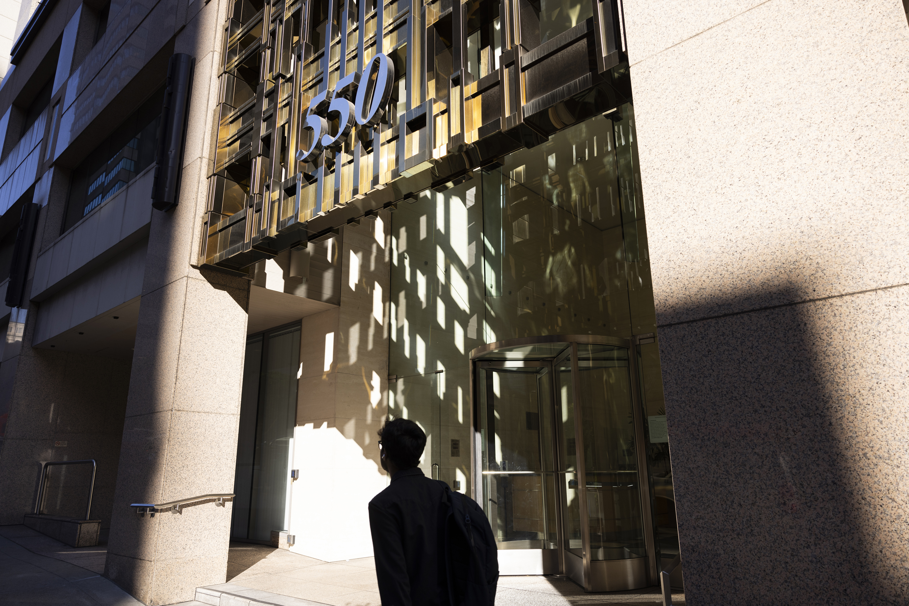 A person stands in front of a modern building marked &quot;550,&quot; with sunlight casting sharp shadows on its glass facade.