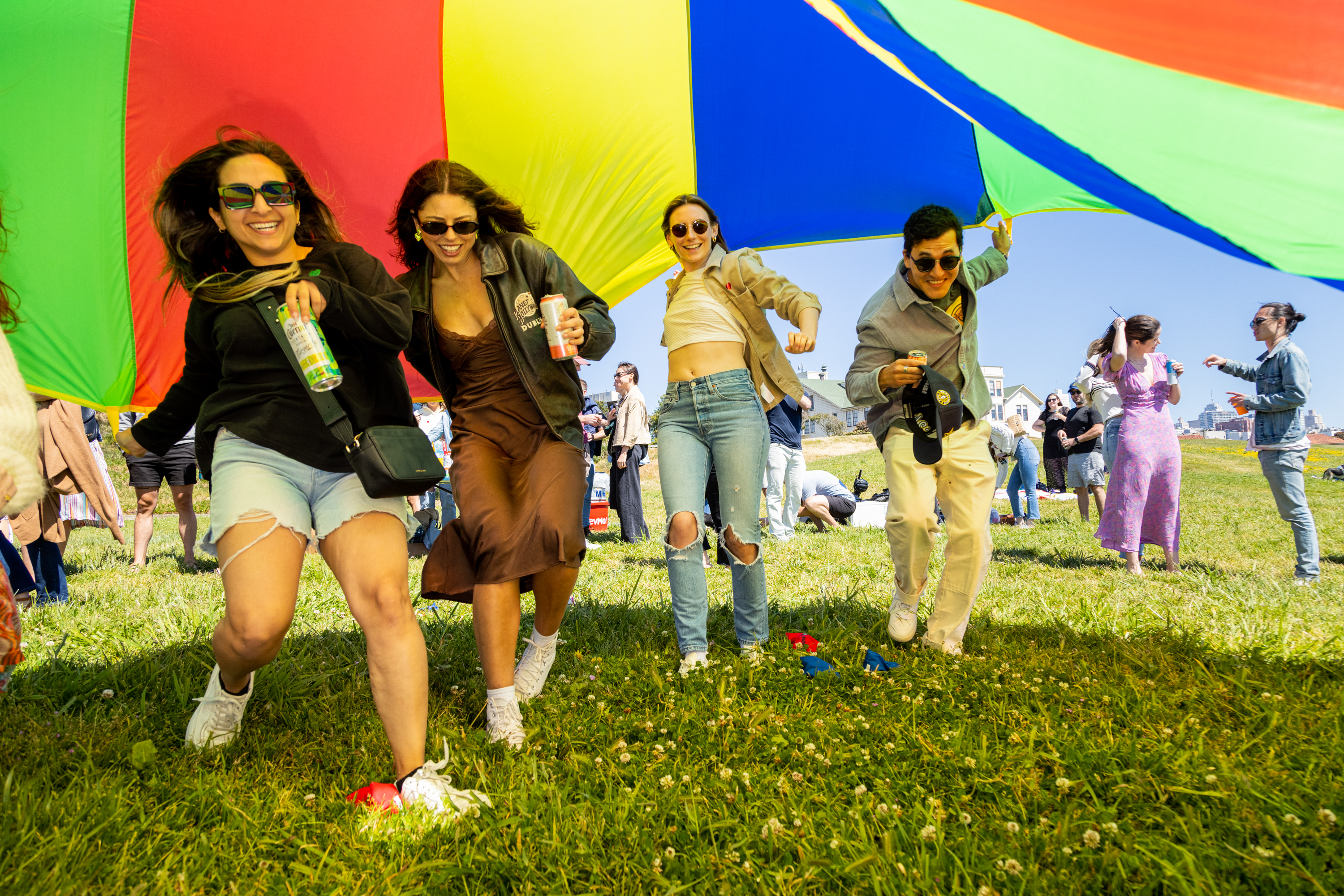 Four joyful individuals run under a colorful, large parachute on a sunny day at a lively outdoor event while holding drinks.