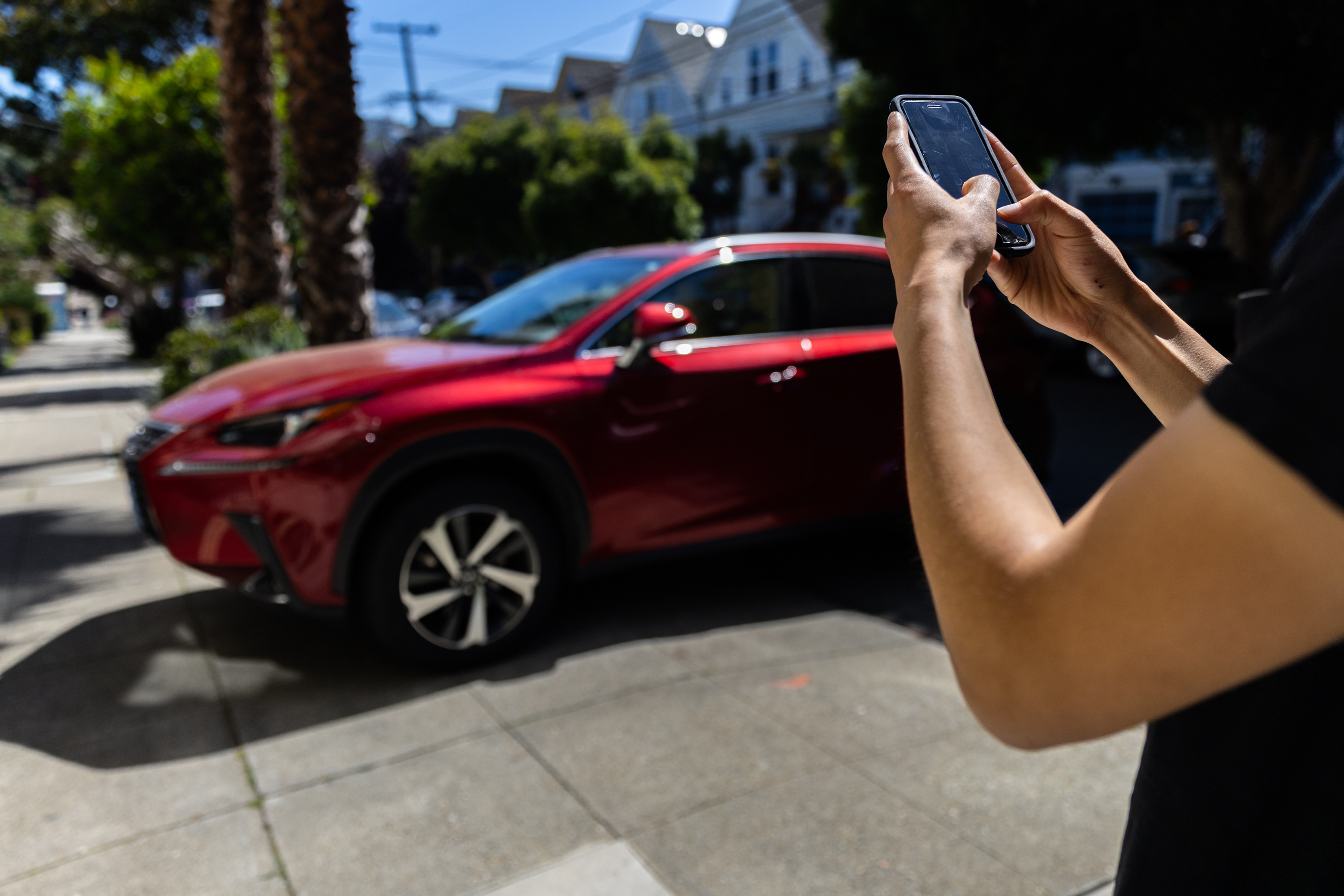 A person is holding a smartphone, focusing on a red car parked on a sunny street lined with trees and houses.