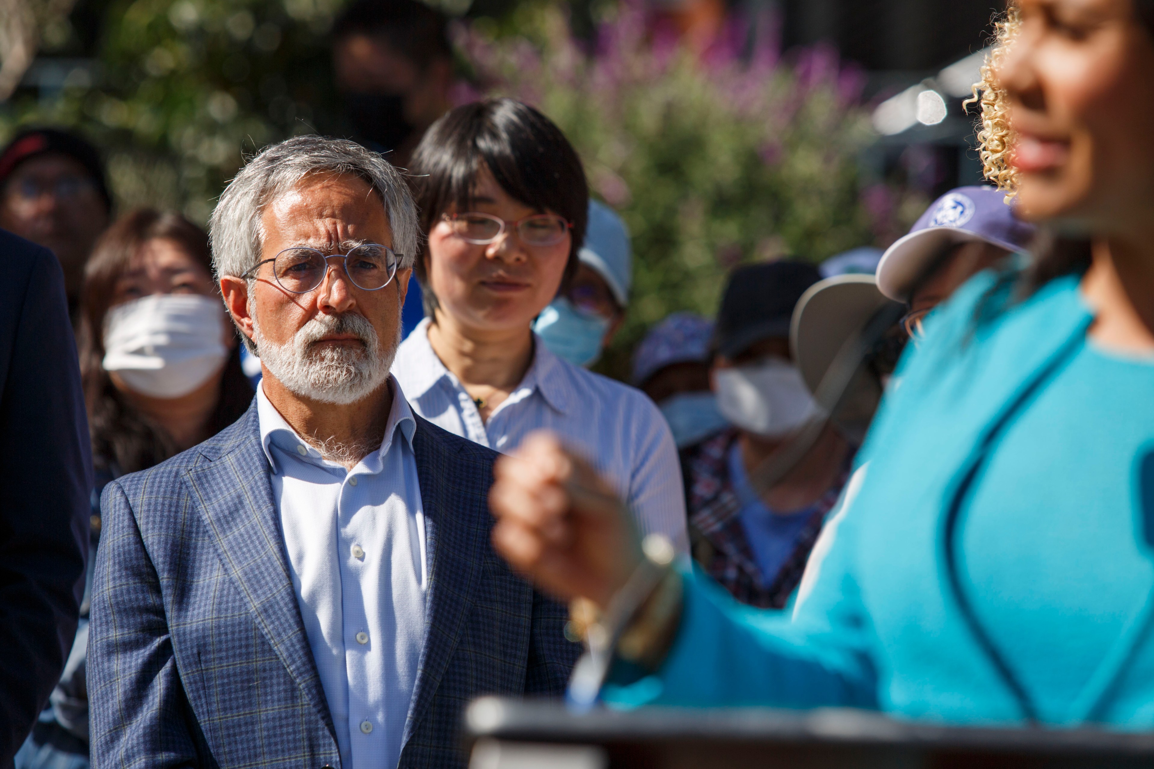 A man in a blue plaid jacket intently listens to a woman in a teal outfit speaking at a podium. Several masked and unmasked people stand in the background.