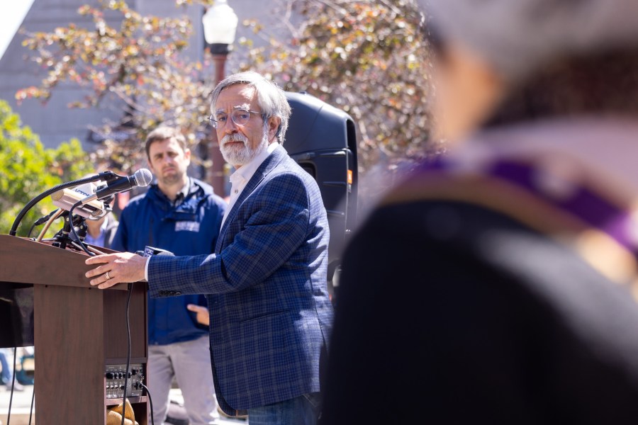 An older man with gray hair and glasses speaks at an outdoor podium. A man in the background watches, and a blurred figure is in the foreground.