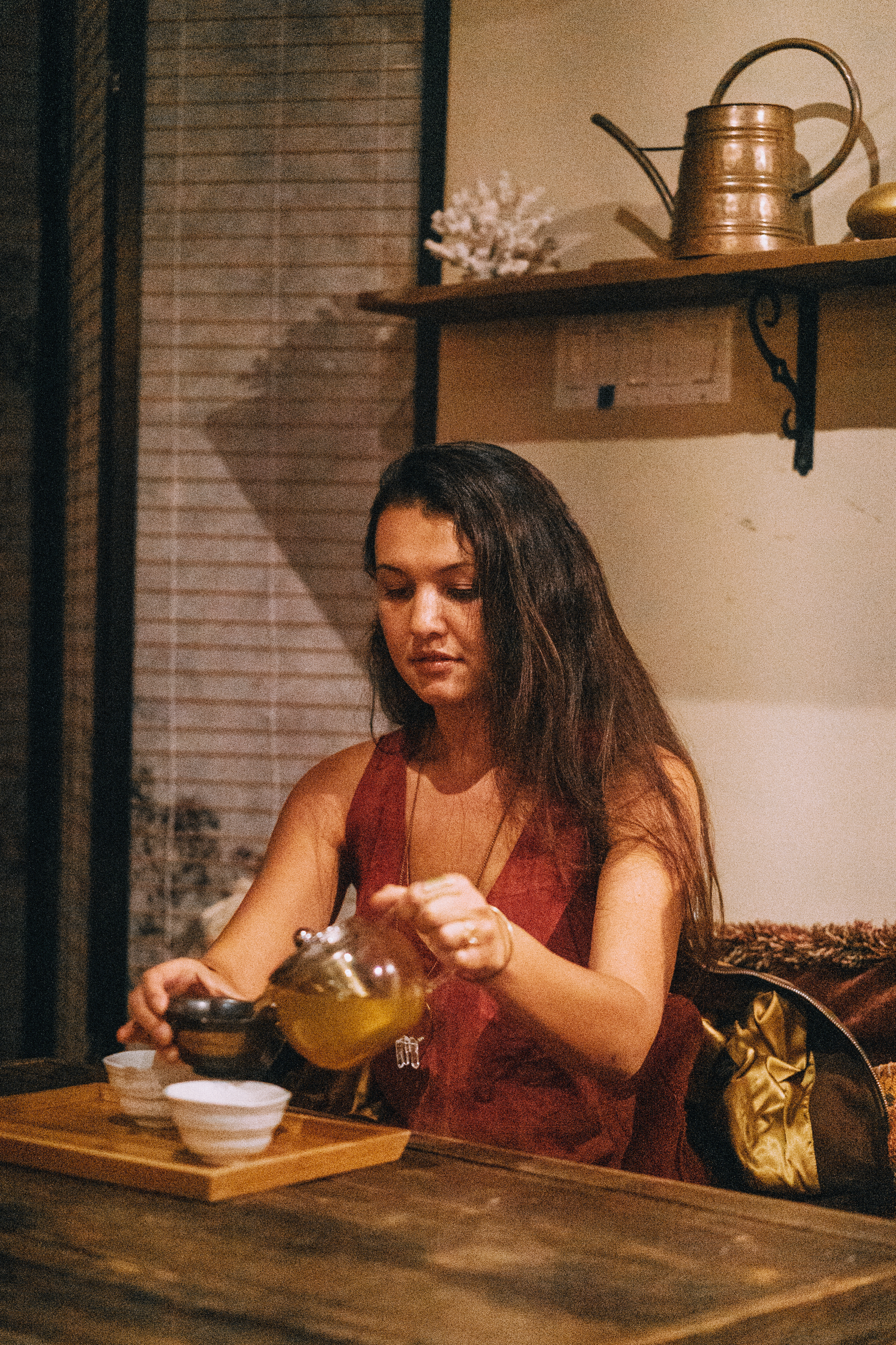 A woman in a red dress pours tea from a glass teapot into white cups on a wooden tray. She is seated at a rustic wooden table in a cozy, dimly lit room.