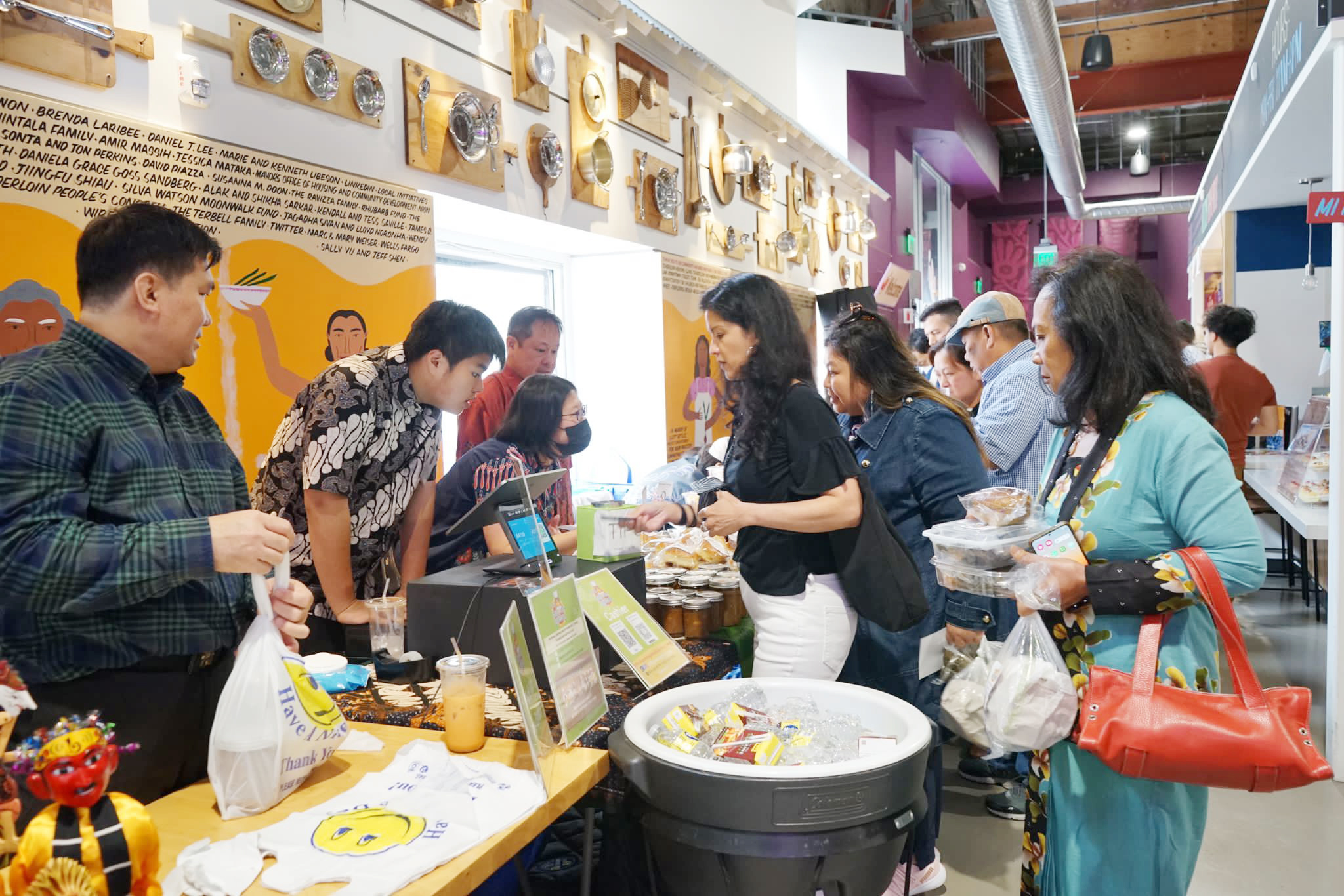 People browse and shop at a crowded indoor market, examining products at a colorful stall decorated with wooden masks.