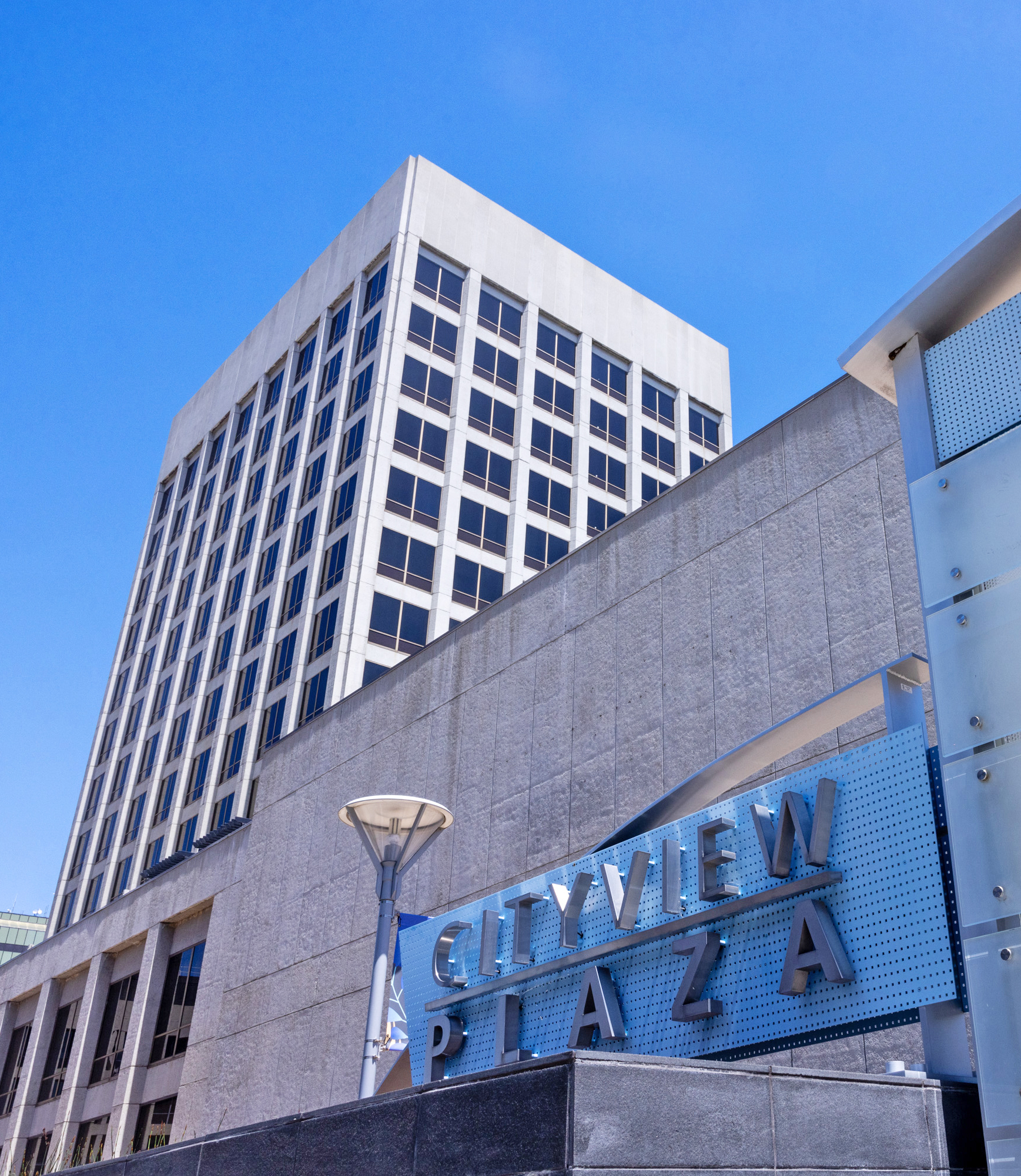 The image shows a tall, modern office building with multiple windows. In the foreground, there's a sign that reads &quot;Cityview Plaza&quot; against a clear blue sky.