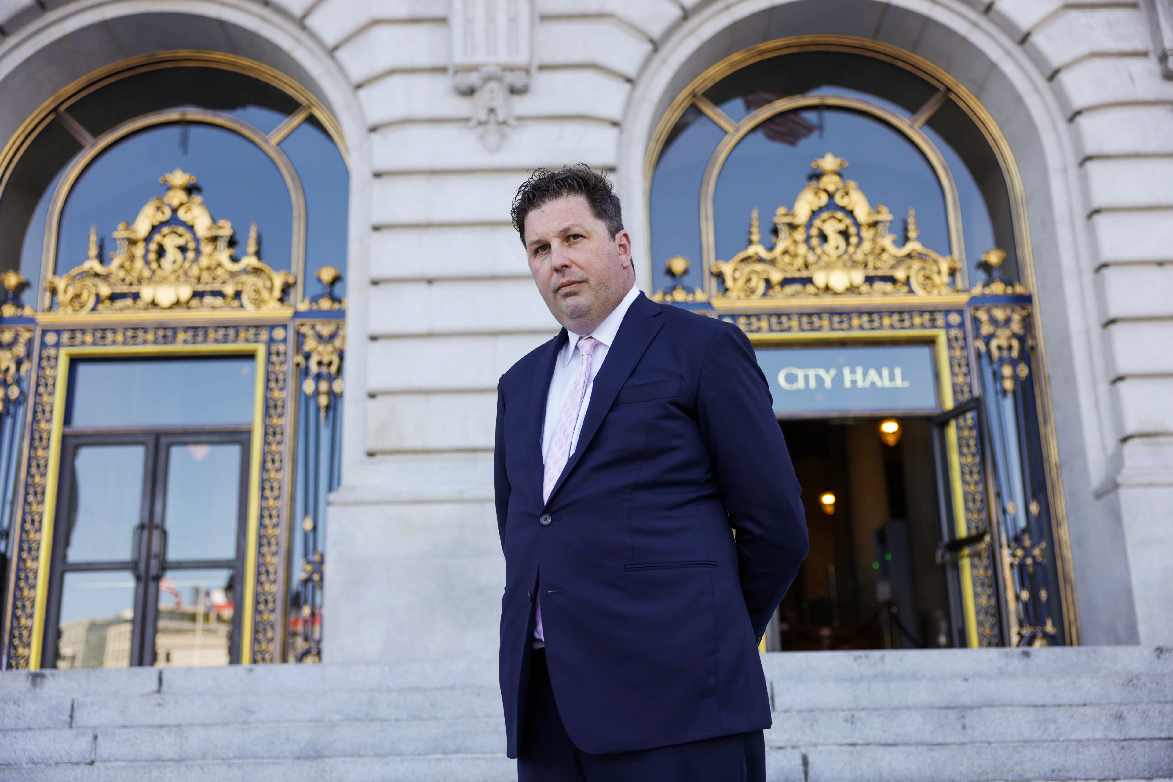 A man in a suit stands in front of an ornate &quot;City Hall&quot; entrance with elegant gold detailing.