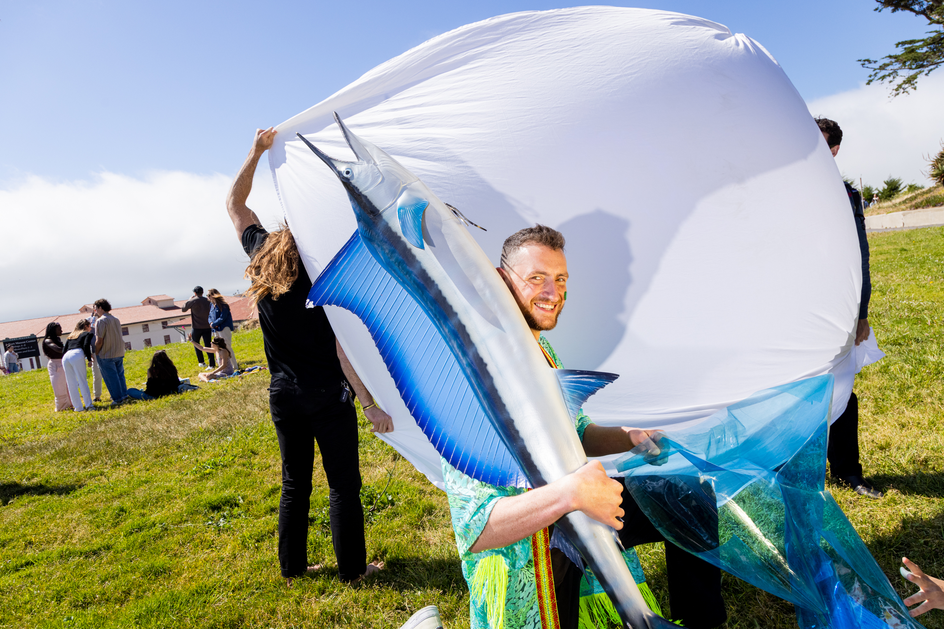 A man smiles while holding a large, sailfish mount at an outdoor event with people and a giant white sheet in the background.
