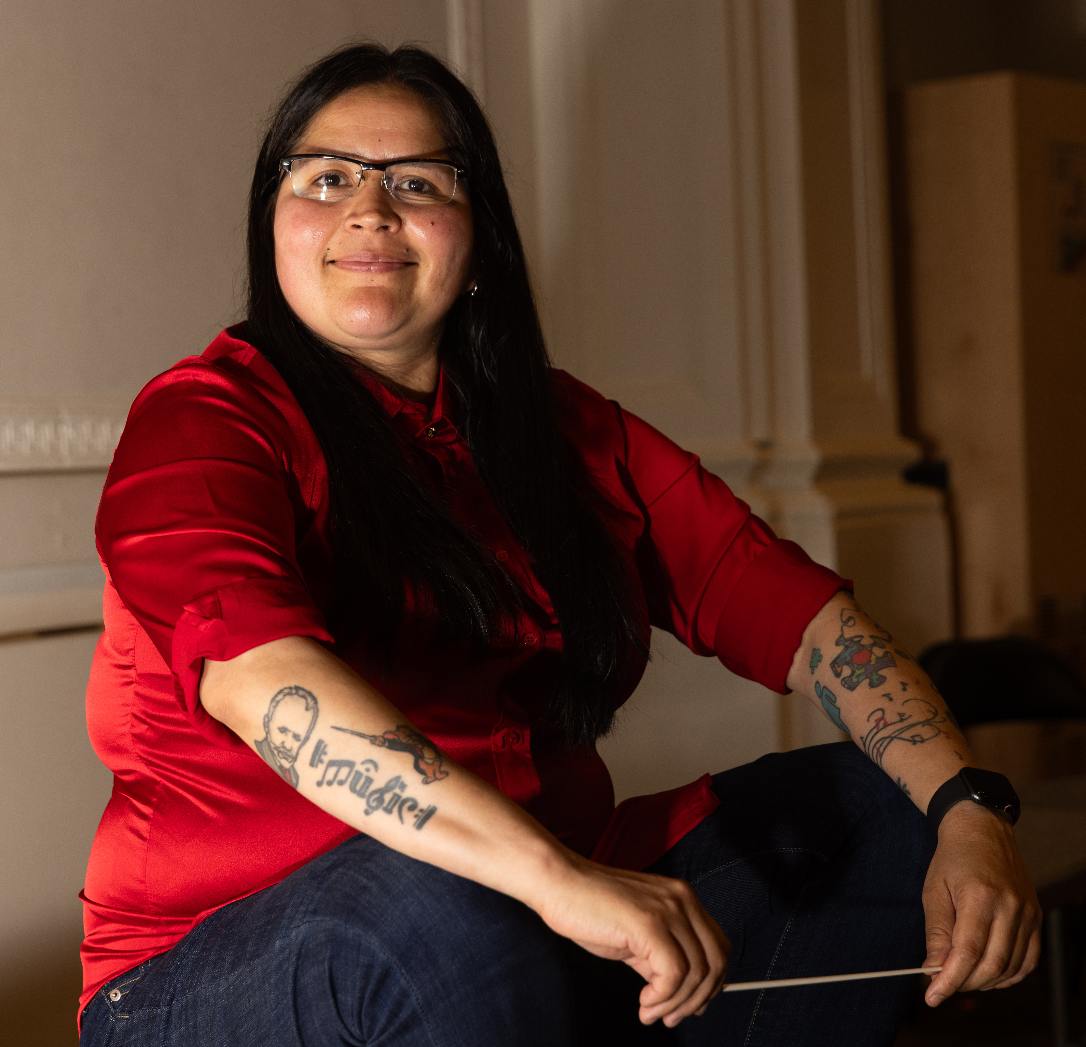 A person with long dark hair and glasses is seated, wearing a red shirt and blue jeans. They have visible tattoos on both forearms and are holding a conductor’s baton.