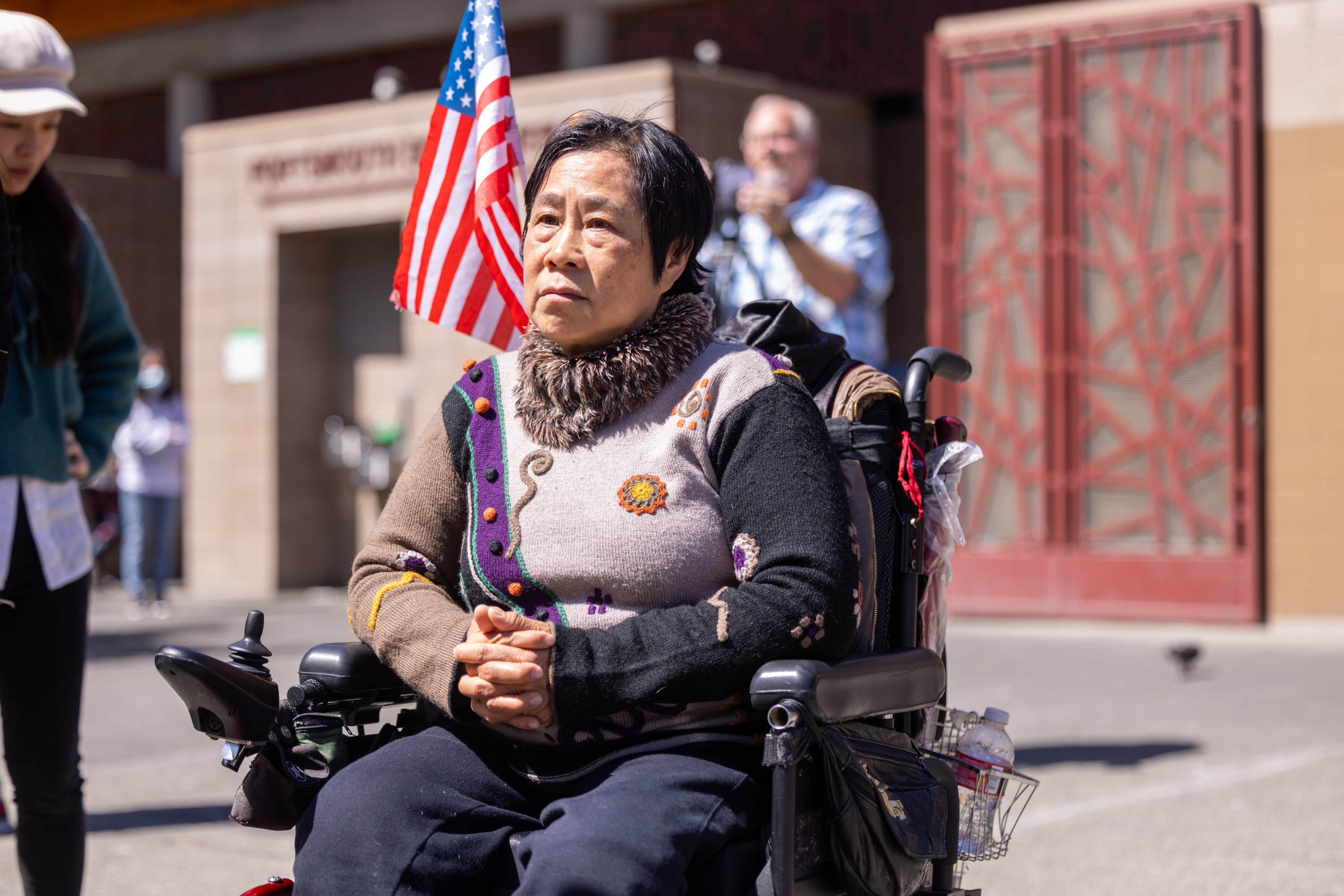 A woman in a colorful sweater sits in a wheelchair displaying an American flag. She is outdoors, flanked by a woman on the left and a man in the background.