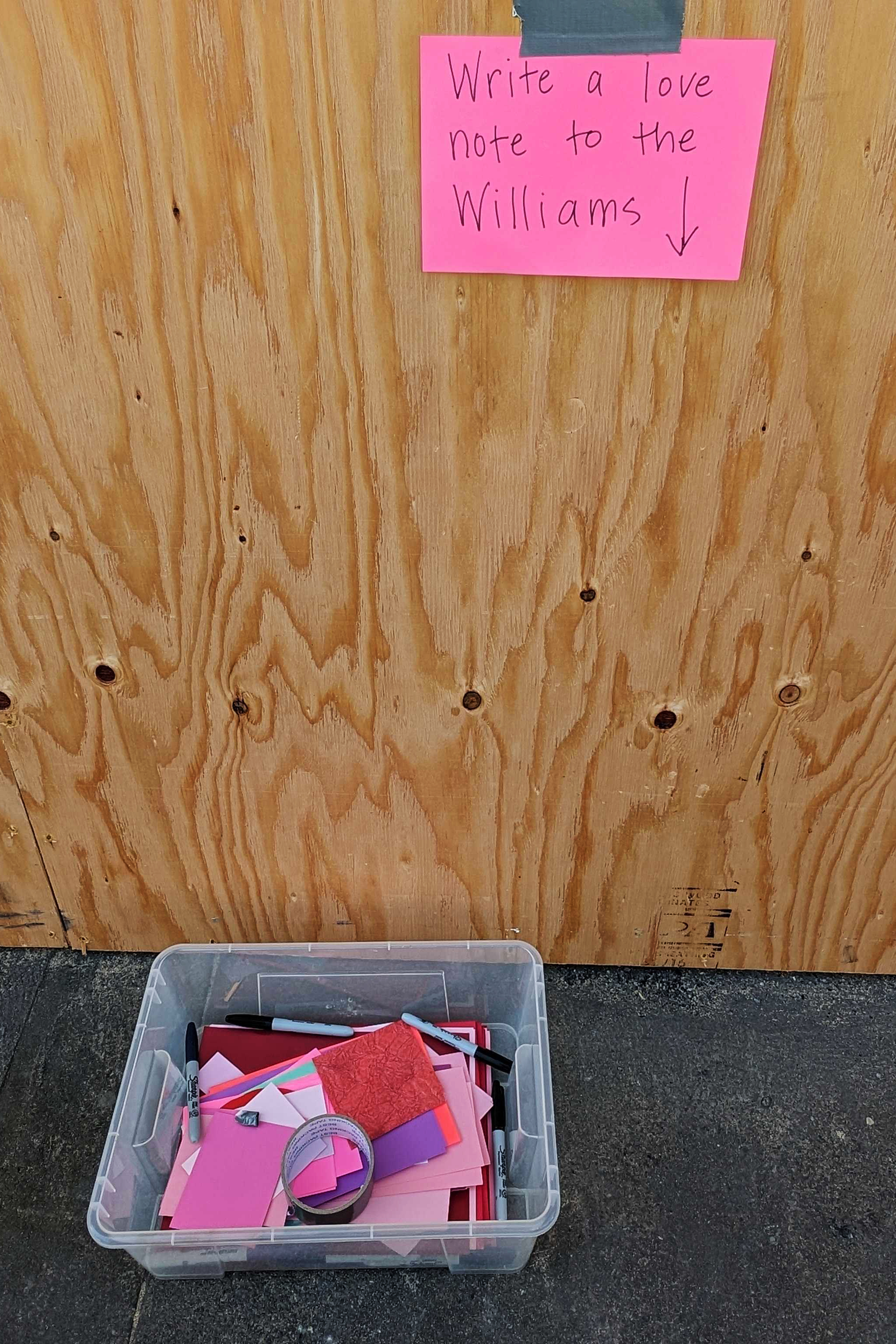 A pink sign on wood reads &quot;Write a love note to the Williams&quot; with an arrow pointing to a bin below, containing markers, tape, and various colored paper.