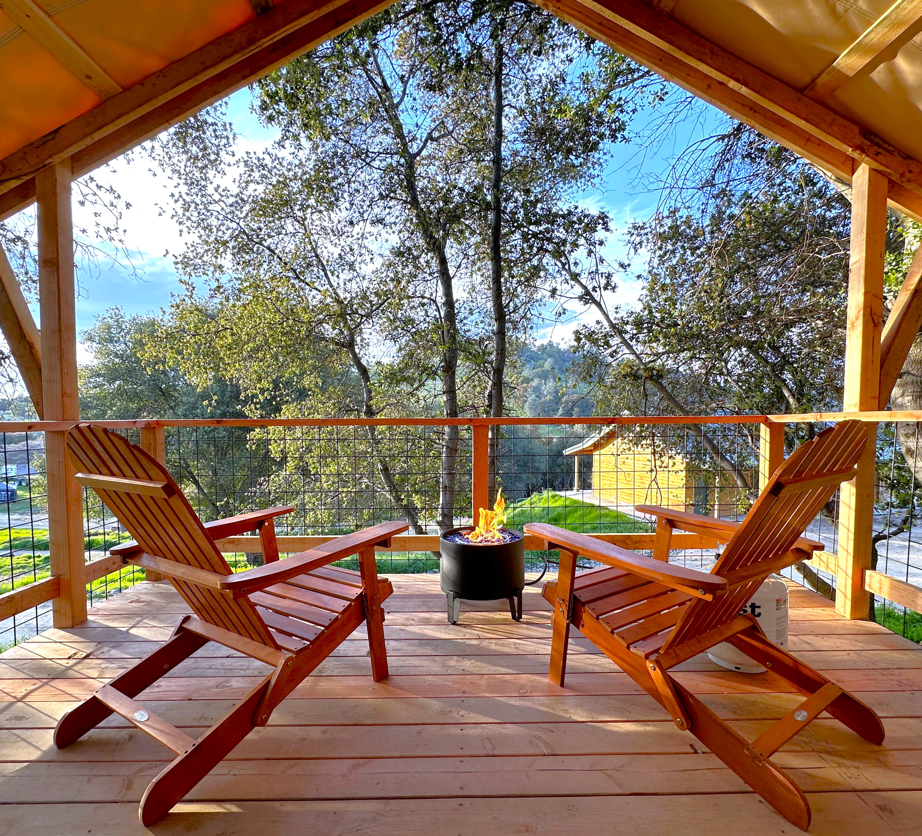 A wooden deck features two reclining chairs facing a small fire pit. Behind is a view of trees and a distant valley, framed by a rustic, open wooden structure.