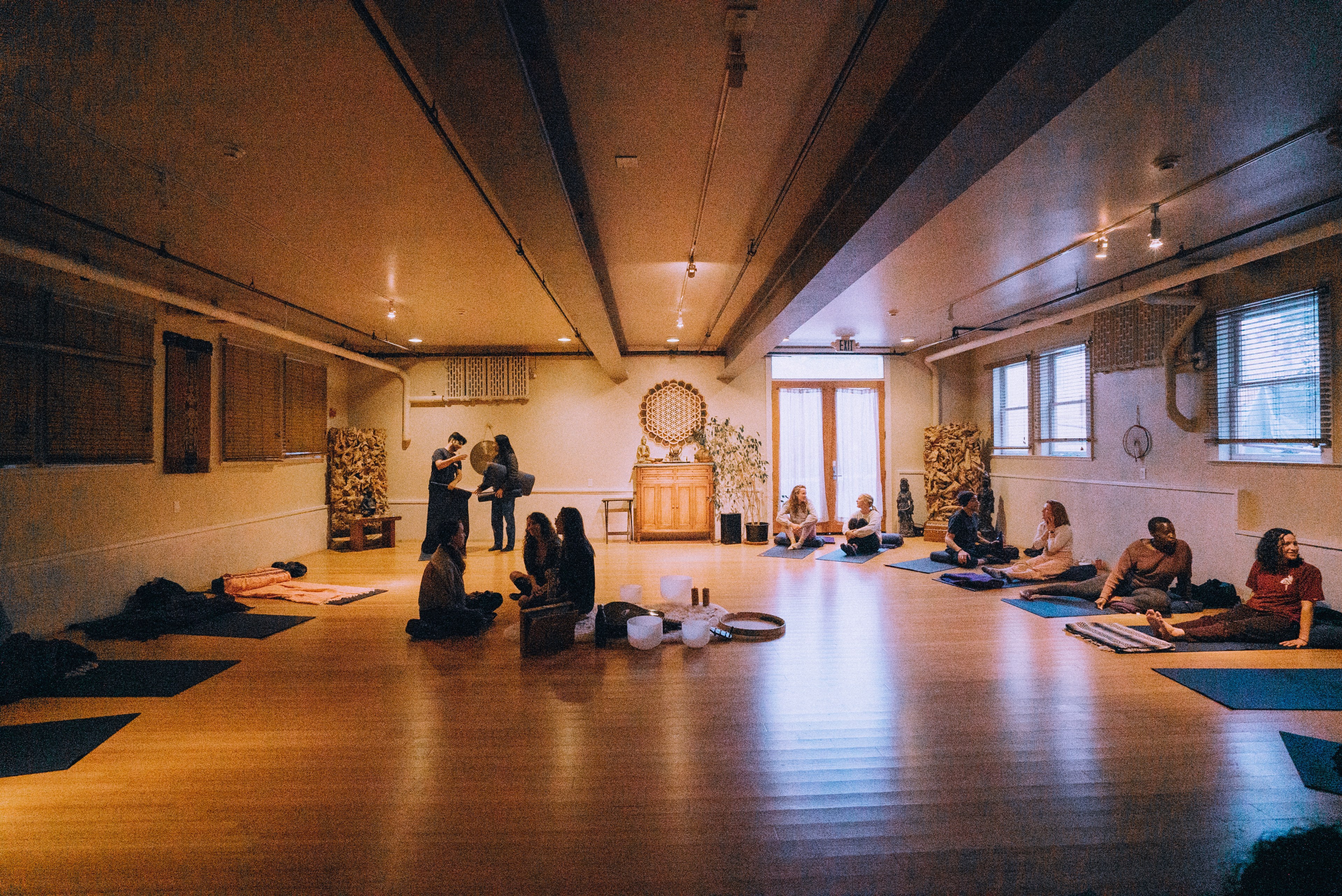 A group of people sit on mats in a serene, well-lit room, likely for a yoga or meditation session. A few individuals converse, and instruments are neatly arranged in the center.