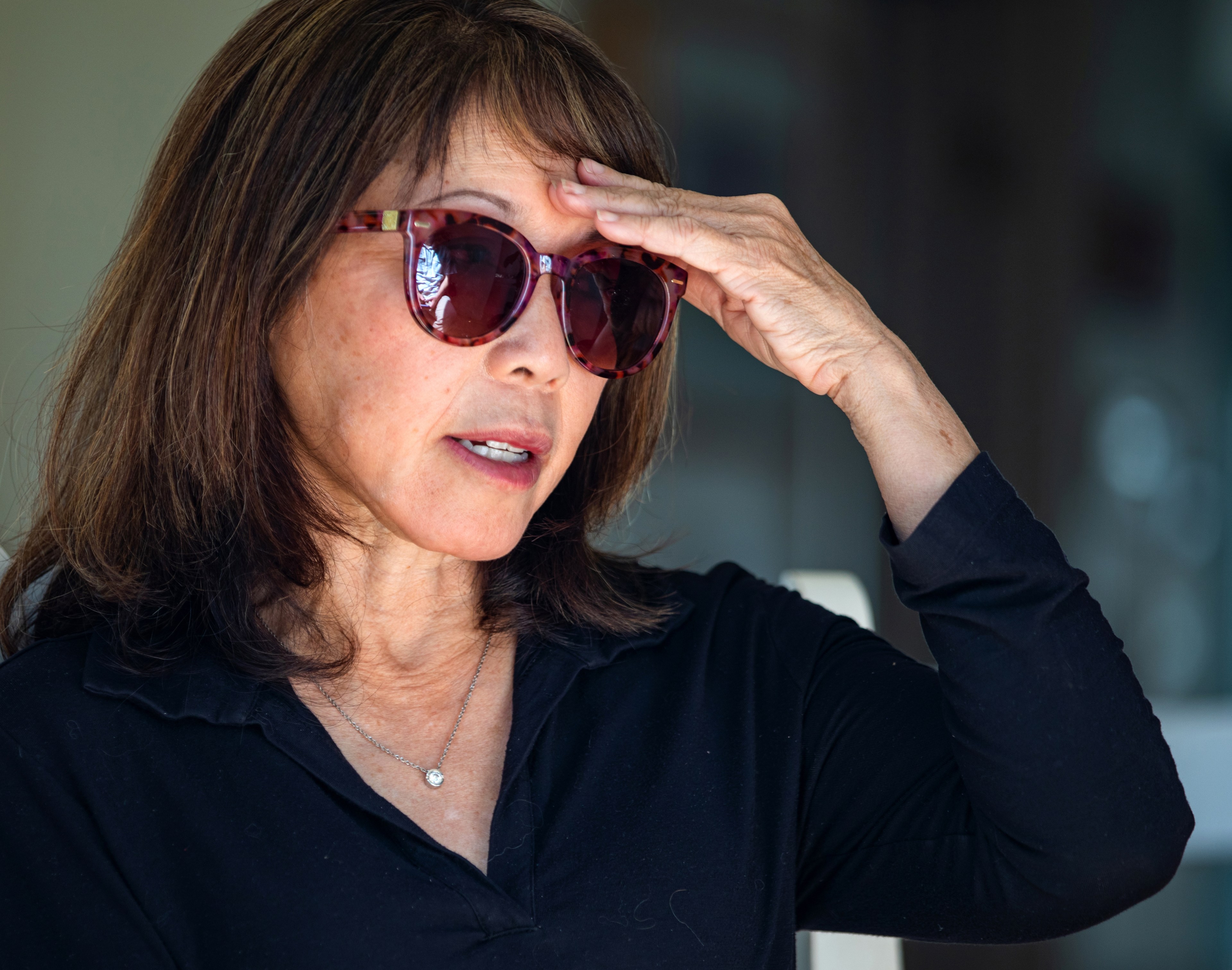 A woman in sunglasses with her hand on her forehead, wearing a black shirt and a necklace.