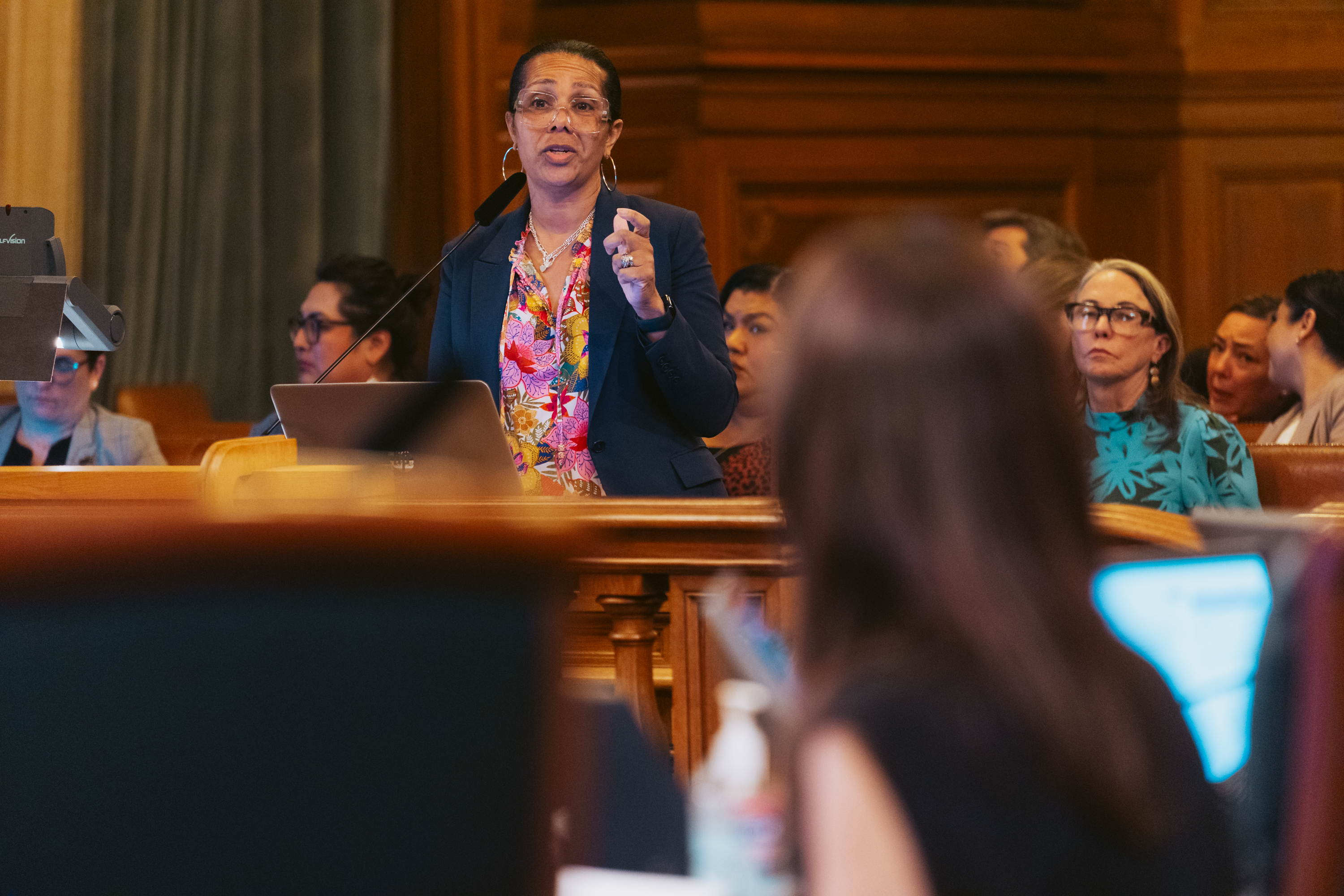 A woman speaks at a podium in a wood-paneled room, wearing glasses and a floral shirt. Several people are seated behind her, attentively listening.