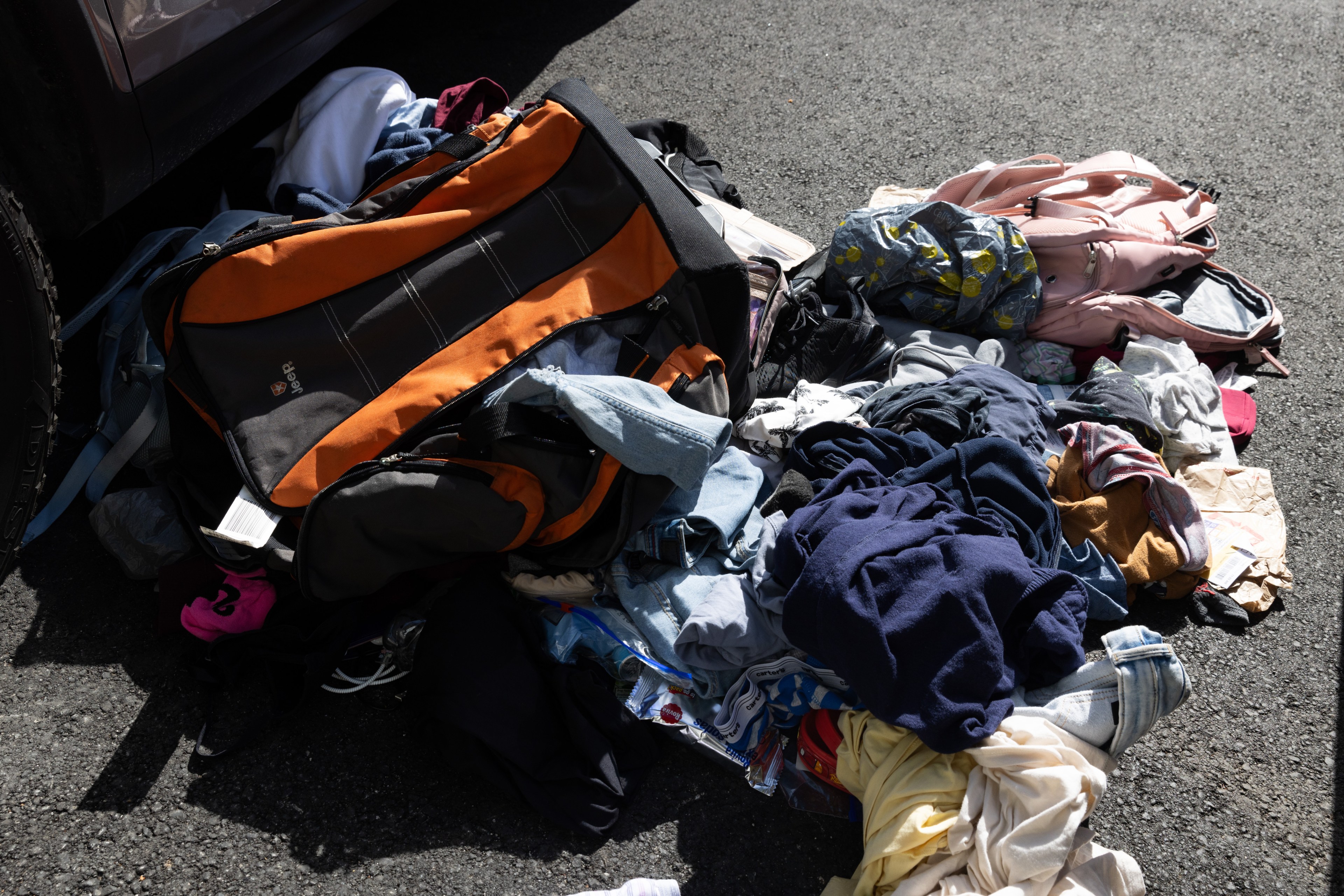 A pile of assorted clothing and several backpacks are strewn on asphalt in a disorganized manner, suggesting a hurried or chaotic situation.
