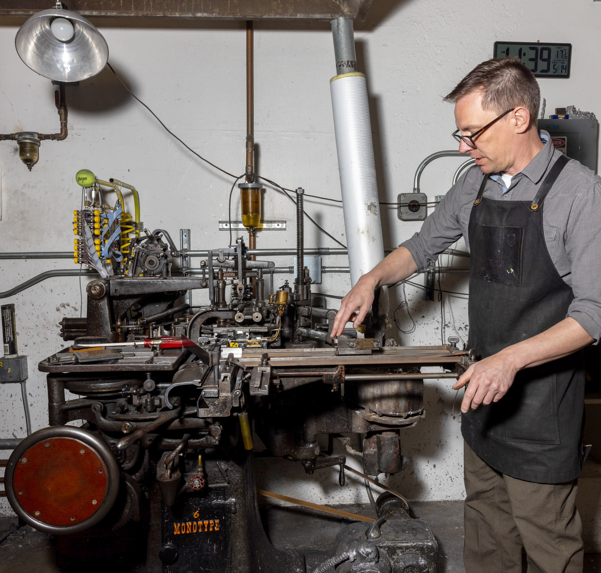 A man in glasses and an apron operates a complex, vintage printing machine in a workshop with tools and pipes around.