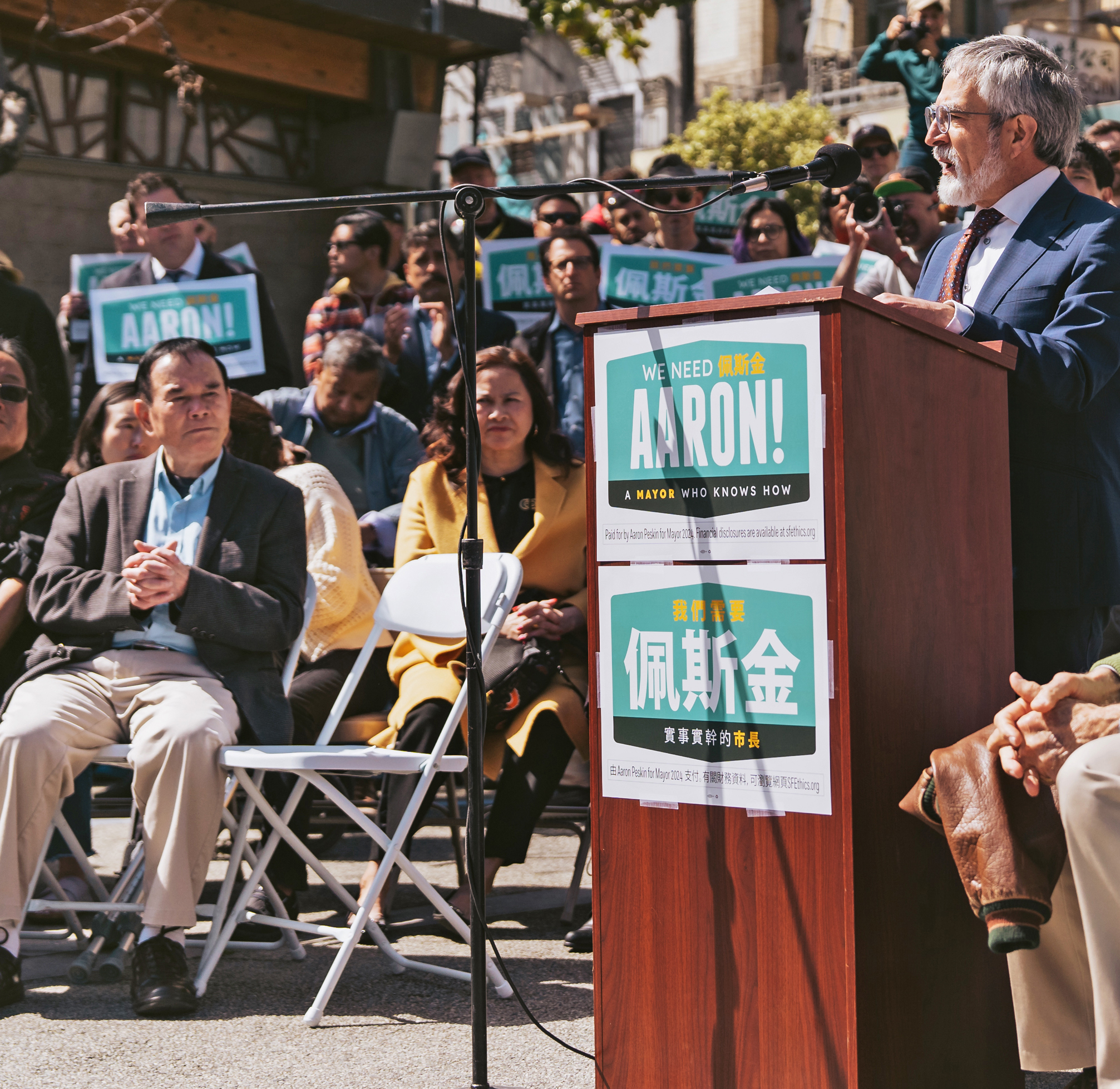 A man speaks at a podium with a &quot;We Need Aaron!&quot; sign, to an attentive audience holding similar signs at an outdoor mayoral campaign event.