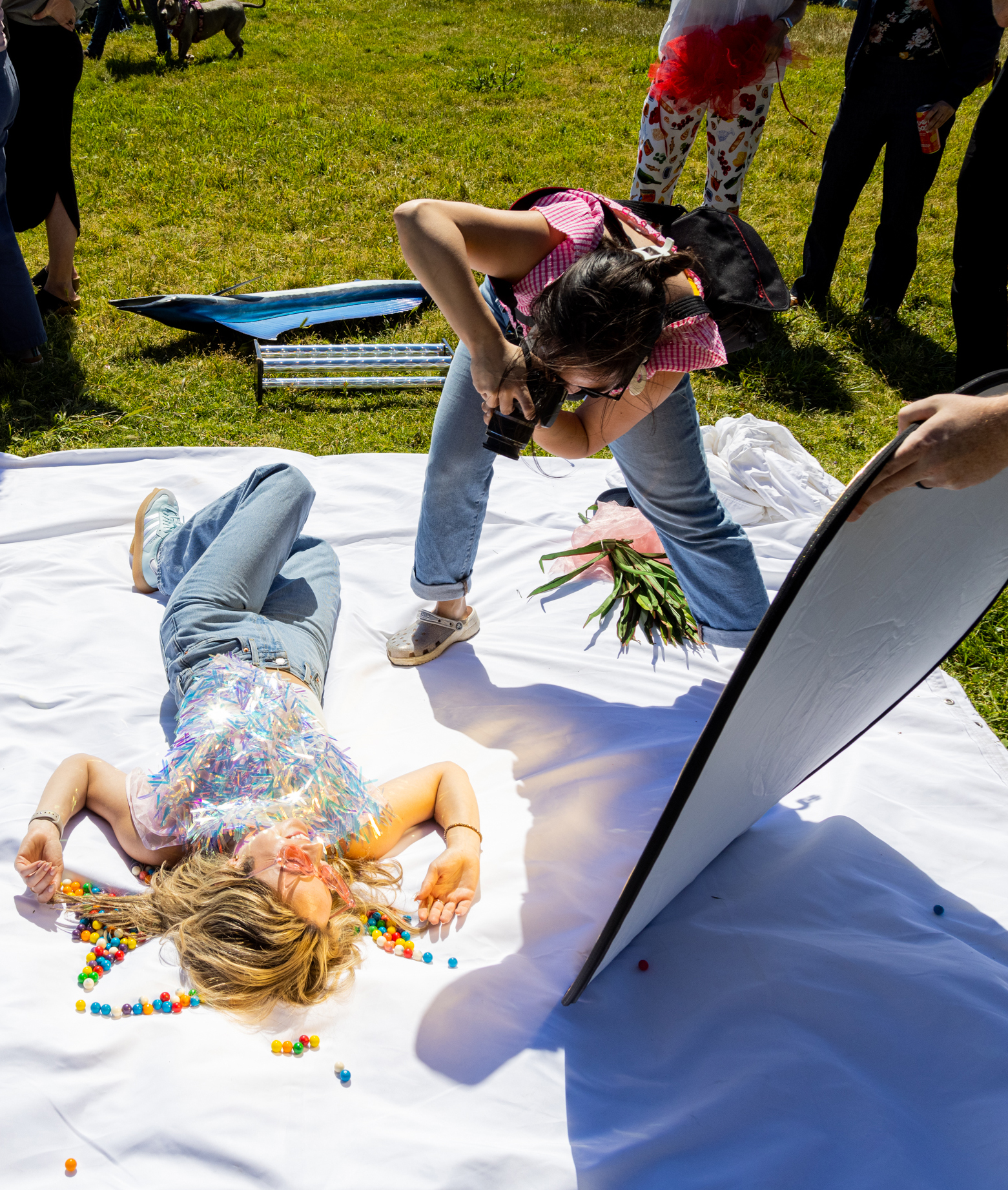 A woman photographs another lying on a white sheet covered in colorful round balls outdoors, with bystanders standing around and a photographer's reflector tilted toward the subject.