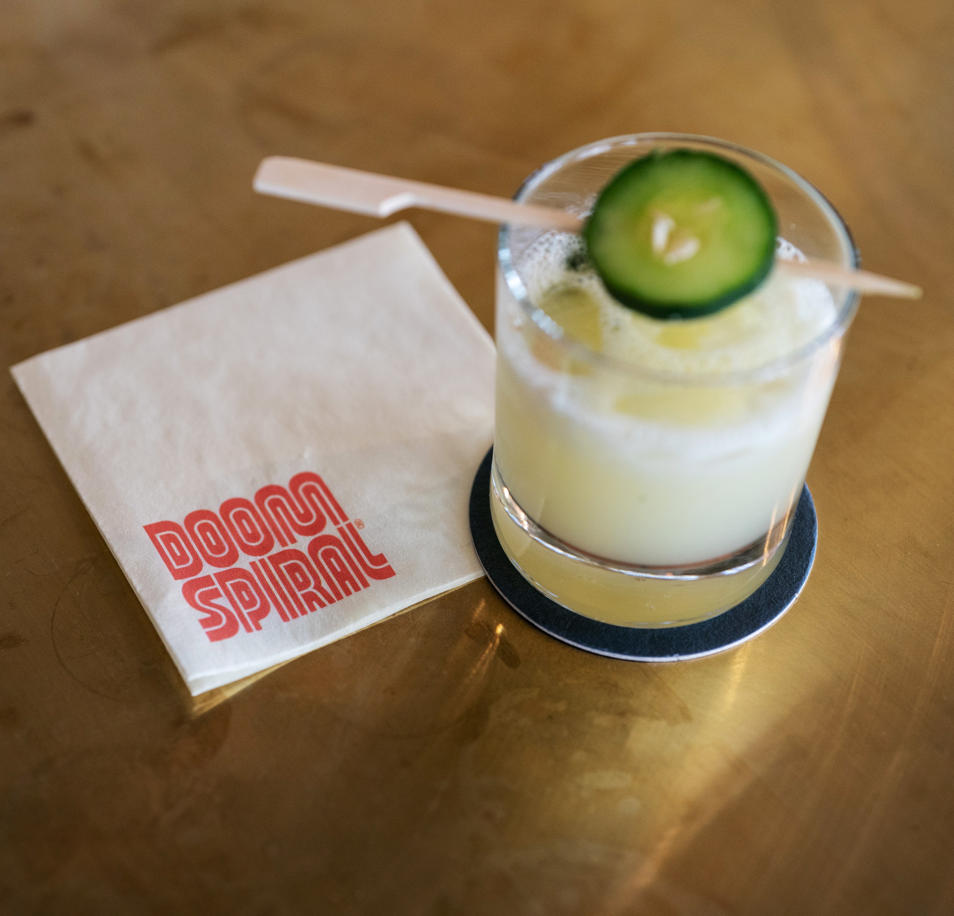 A creamy-looking cocktail with a cucumber slice garnish sits on a dark coaster next to a napkin that reads &quot;DOOM SPIRAL&quot; in red text.