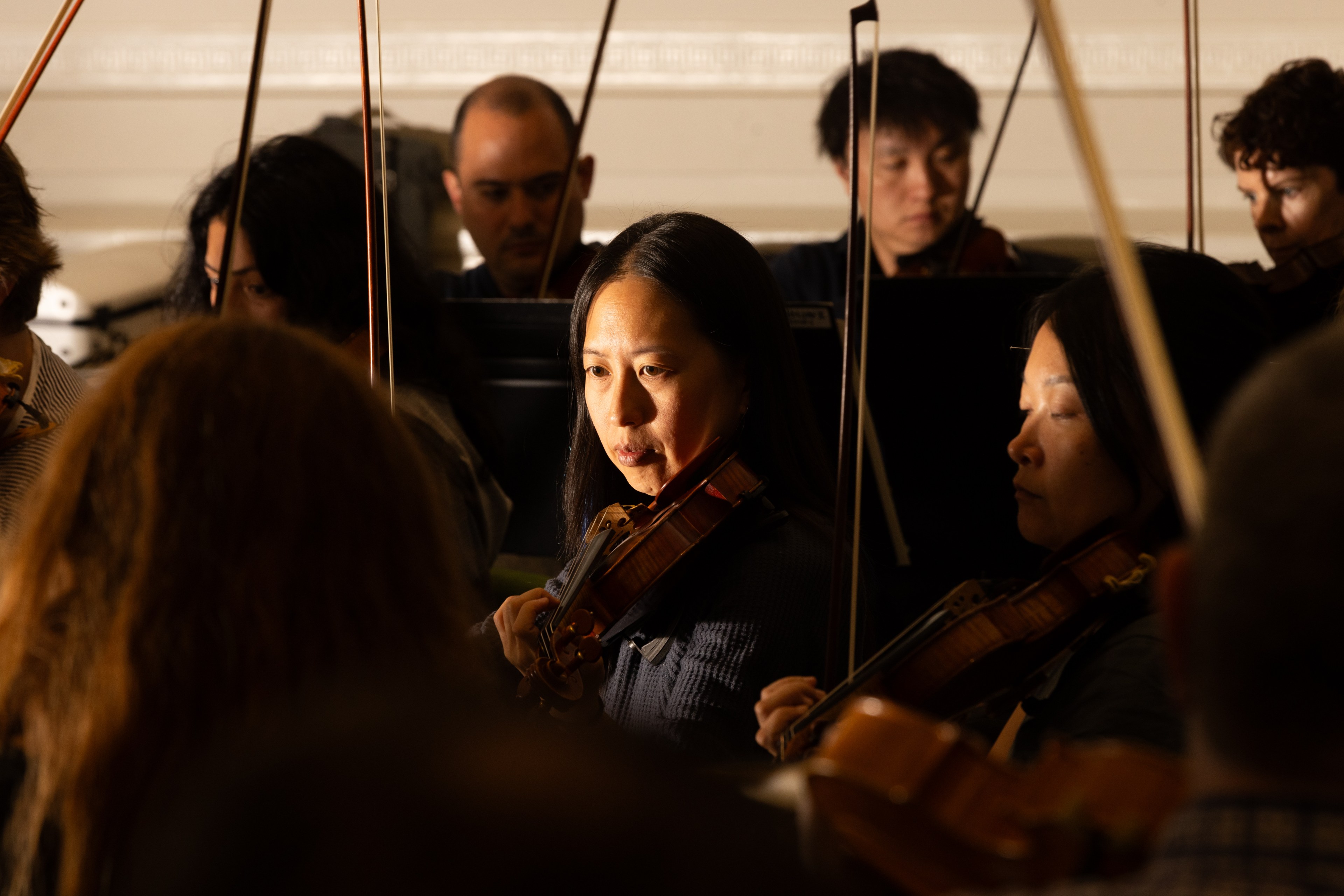 People are playing violins in a dimly lit room, focusing intently on their performance. The spotlight highlights a woman in the foreground.