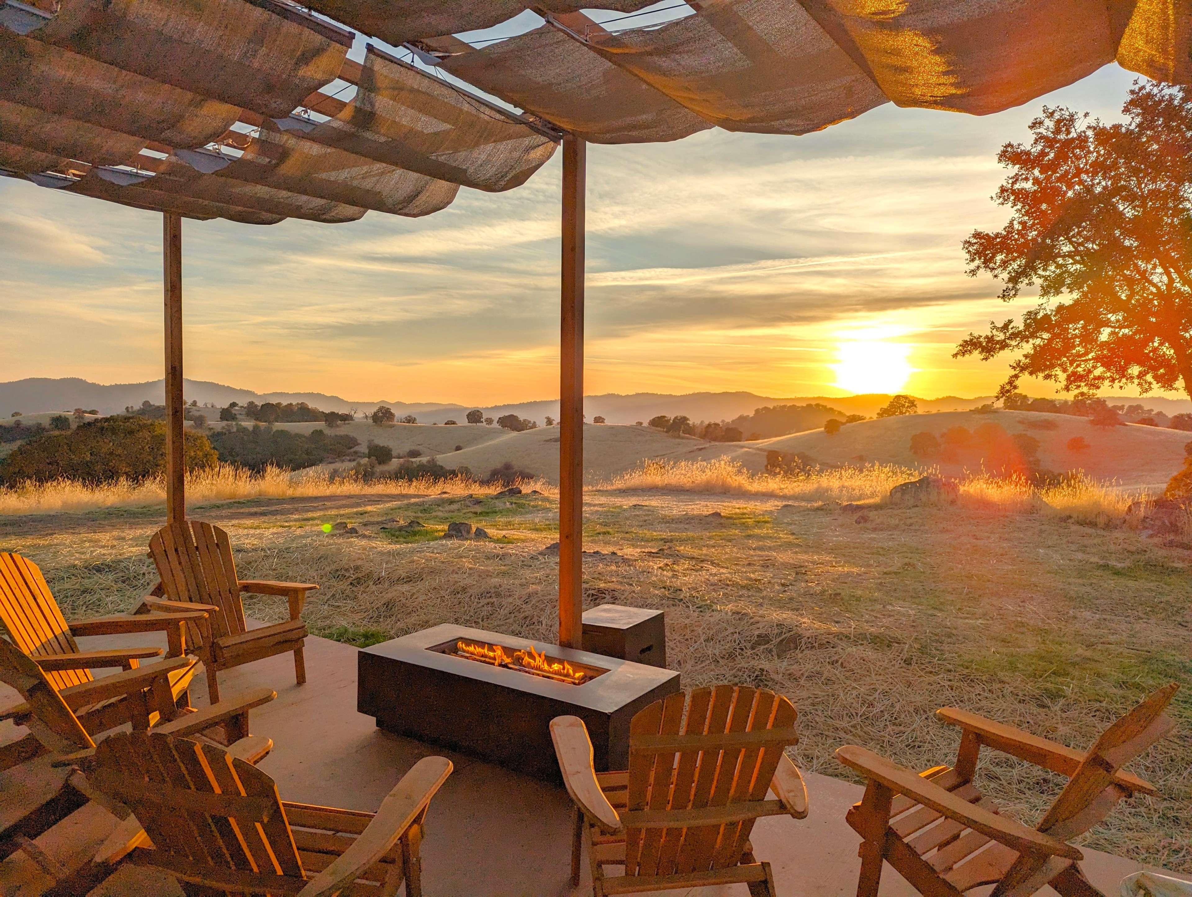 Wooden chairs encircle a fire pit under a canopy, overlooking a serene landscape of rolling hills during a vibrant sunset.